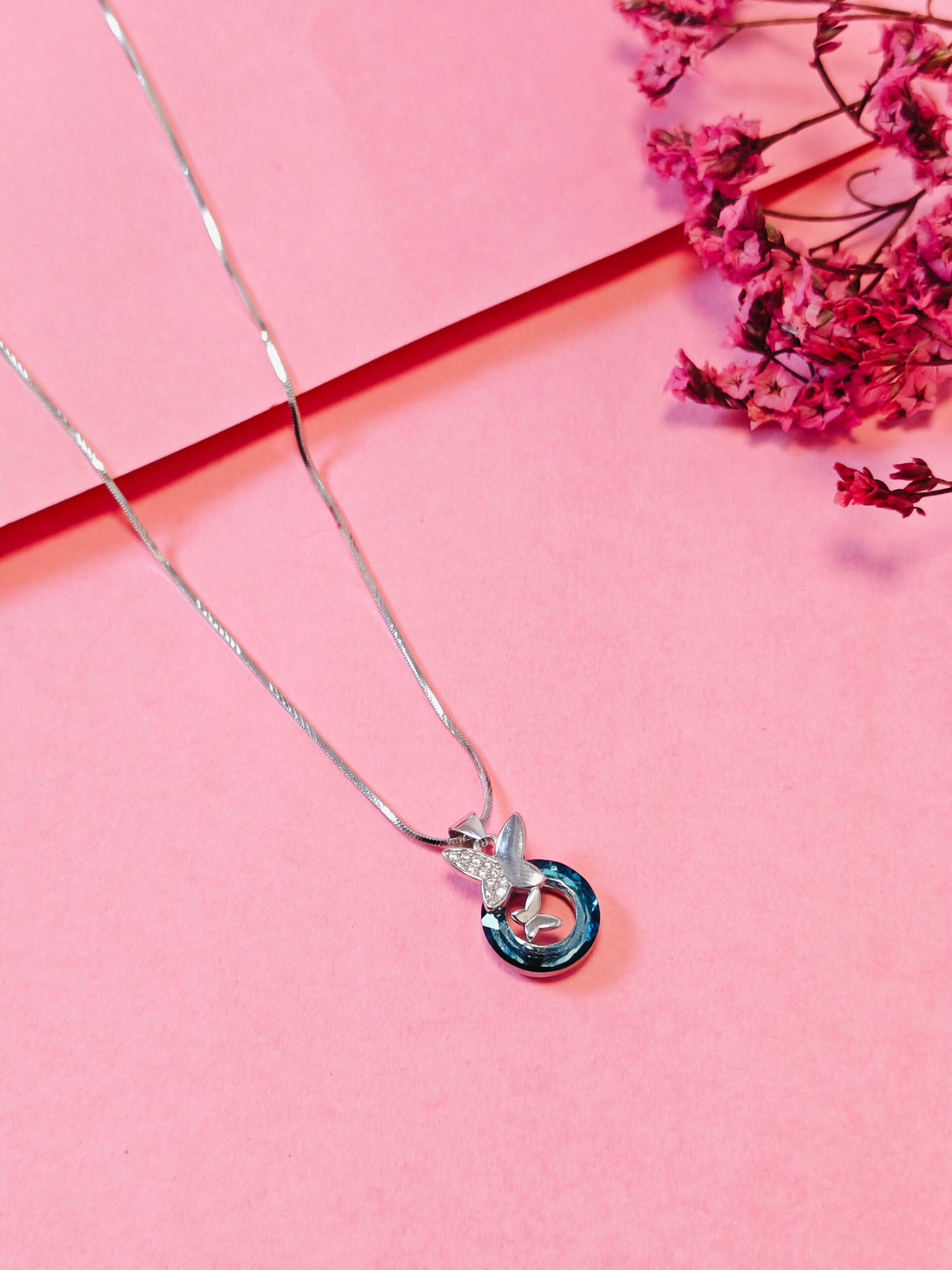 Silver necklace with a blue and clear pendant on a pink background with pink flowers.