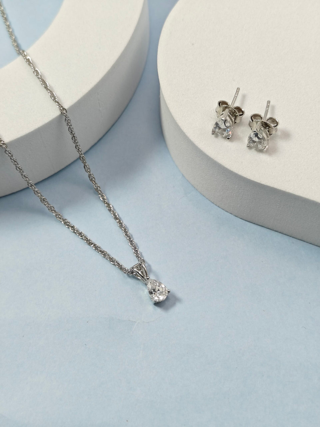 Silver necklace and earrings on a light blue surface with white circular stands.
