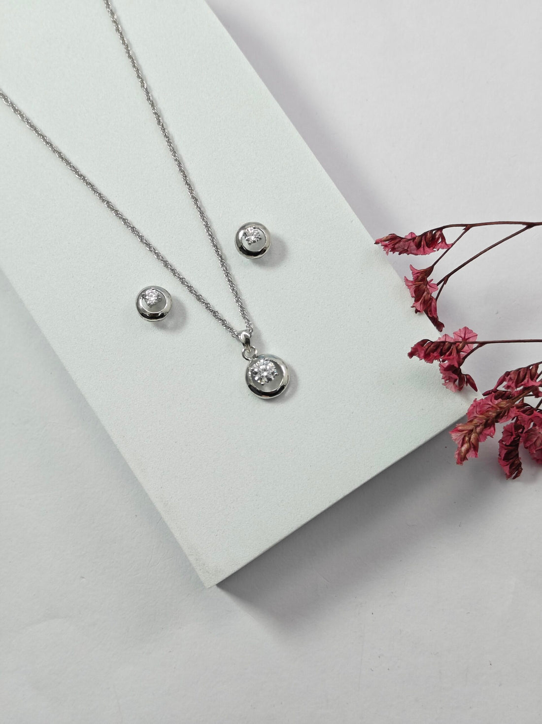 Silver necklace and earrings on a white surface with pink flowers.