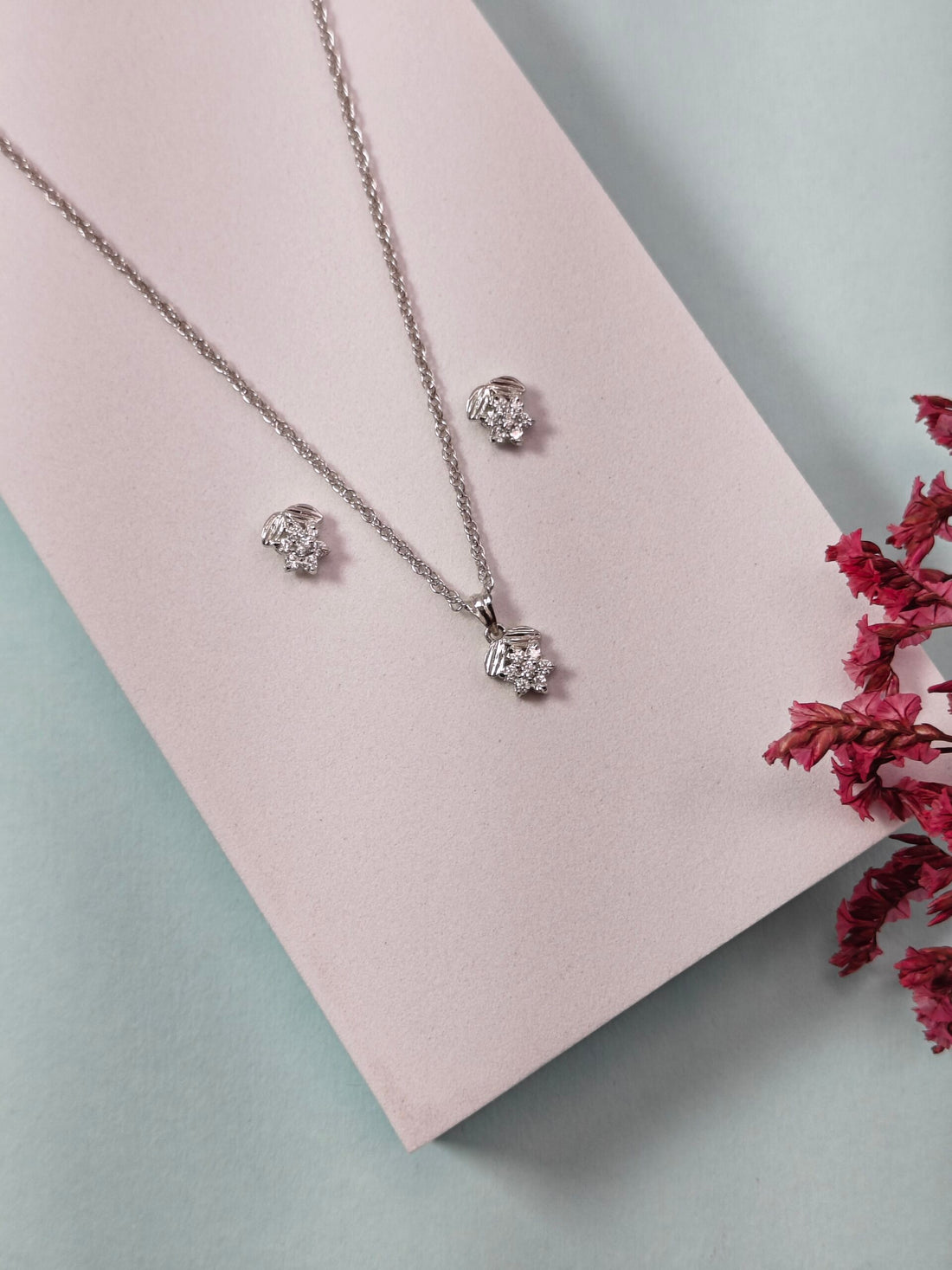 Silver jewelry set with necklace and earrings on a pink cushion, surrounded by red flowers.