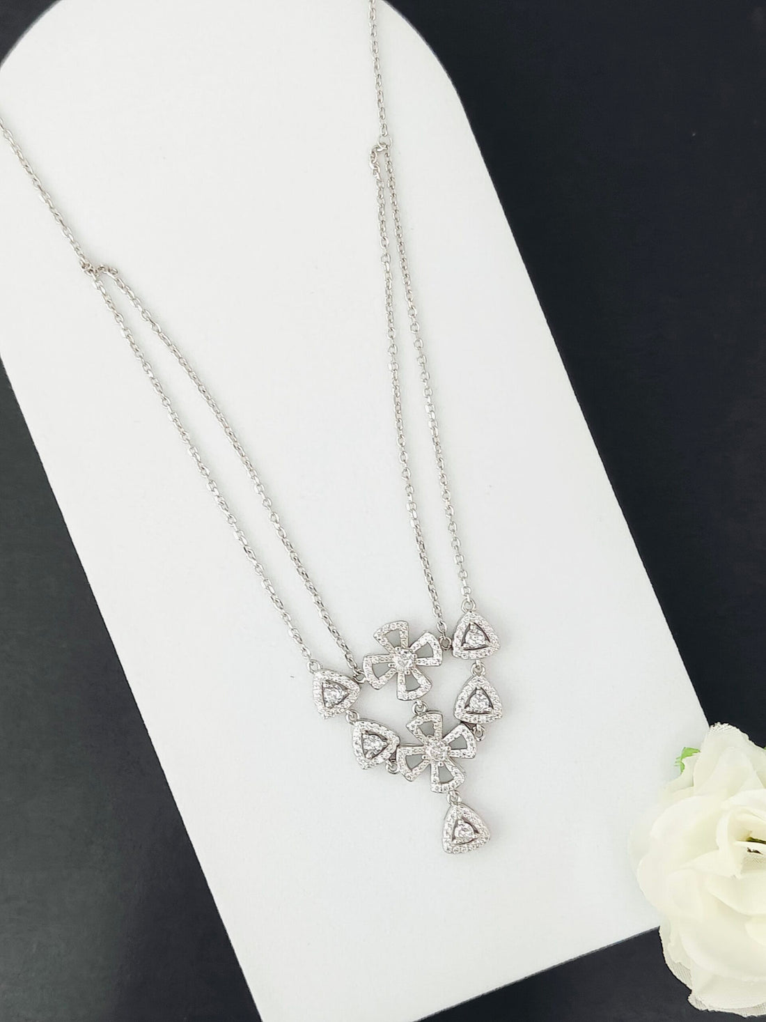 Silver necklace with a heart-shaped pendant on a white stand with a white flower.