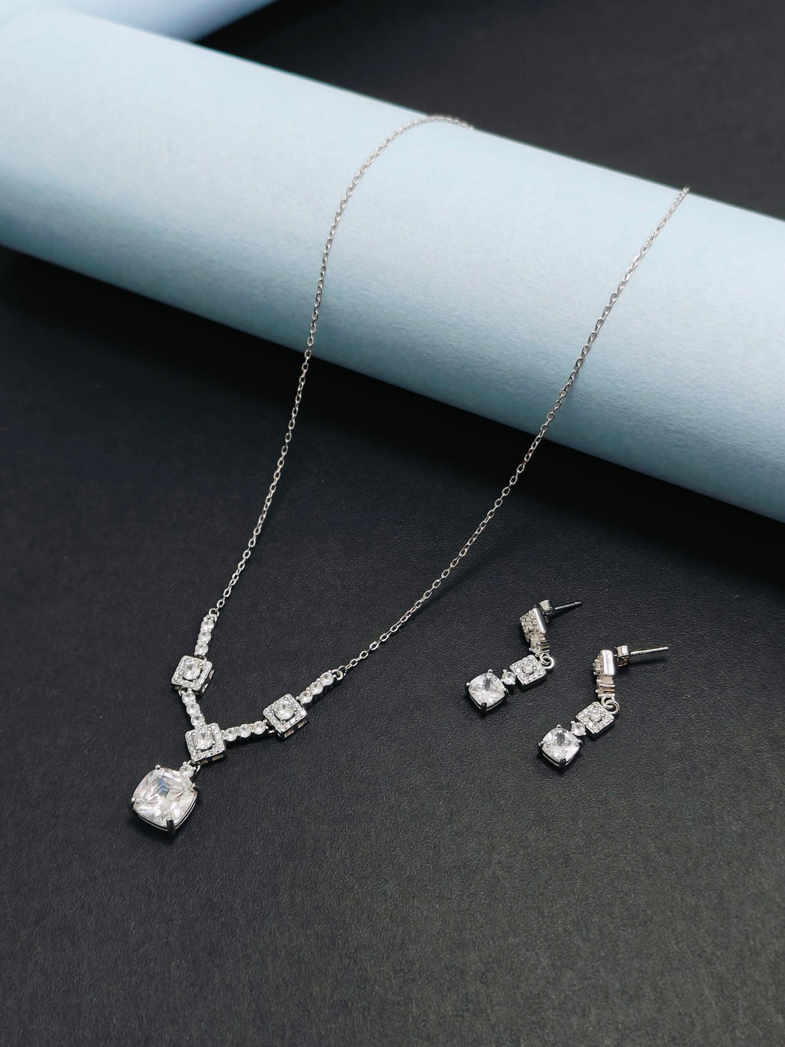 Silver necklace and earrings set on a dark surface with a light blue cylindrical object.