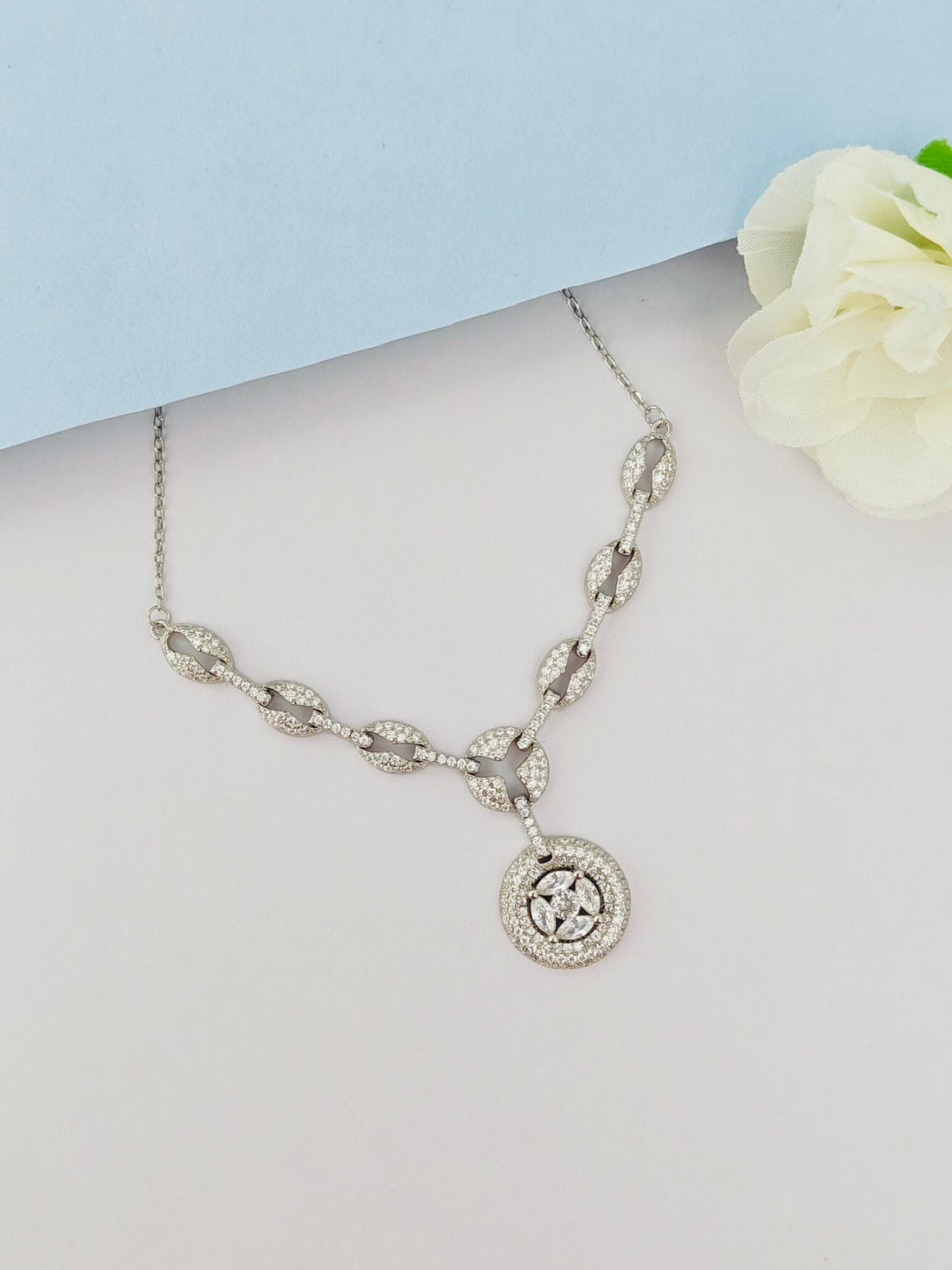 Silver necklace with a circular pendant on a light gray surface with a white flower.