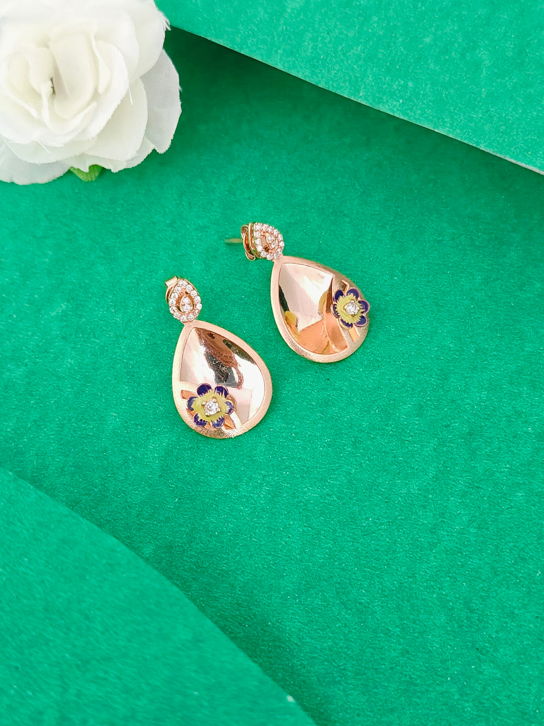Rose gold teardrop earrings with floral details on a green surface with a white flower.