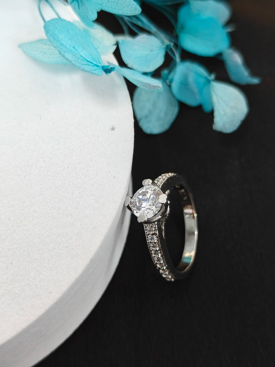 Silver ring with a diamond on a dark surface with blue flowers in the background