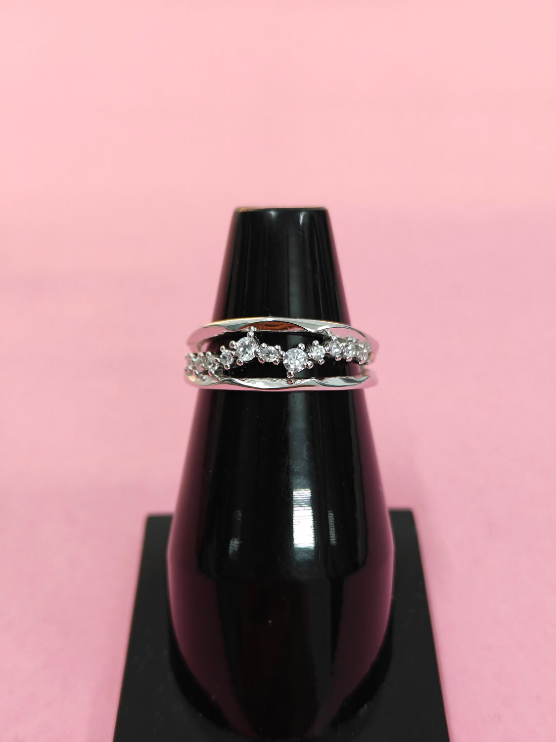 Silver ring with diamonds on a black stand against a pink background