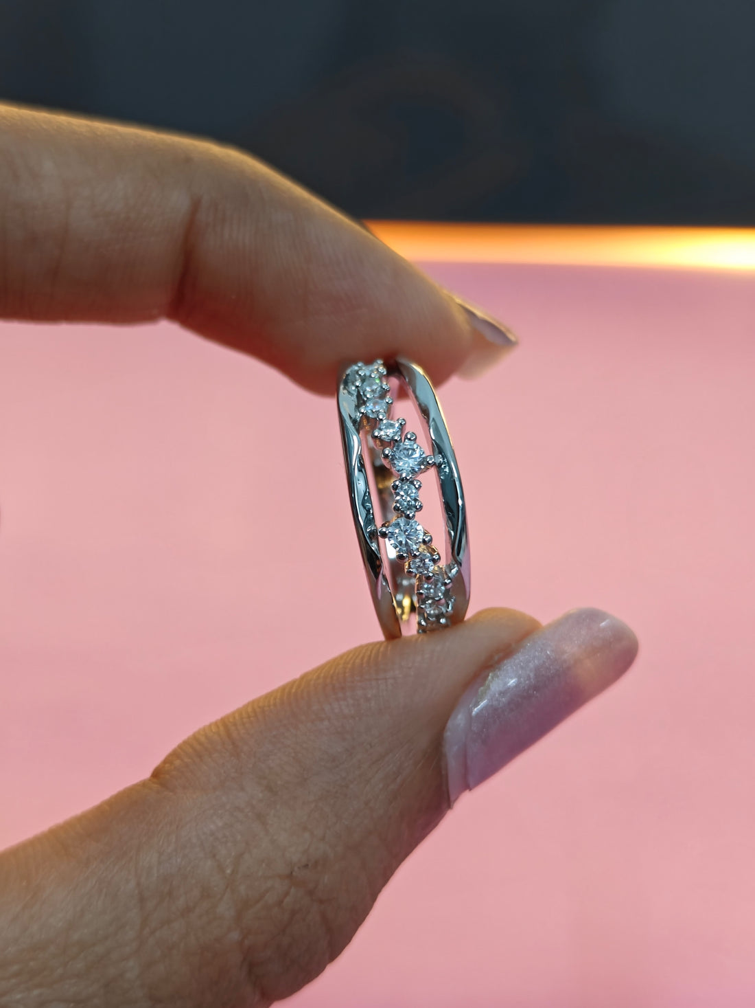 Silver ring with diamonds held between fingers against a pink background
