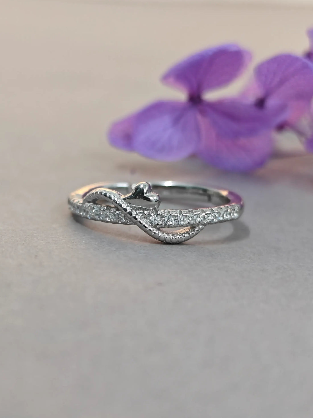 Silver ring with diamond accents on a gray surface with purple flowers in the background