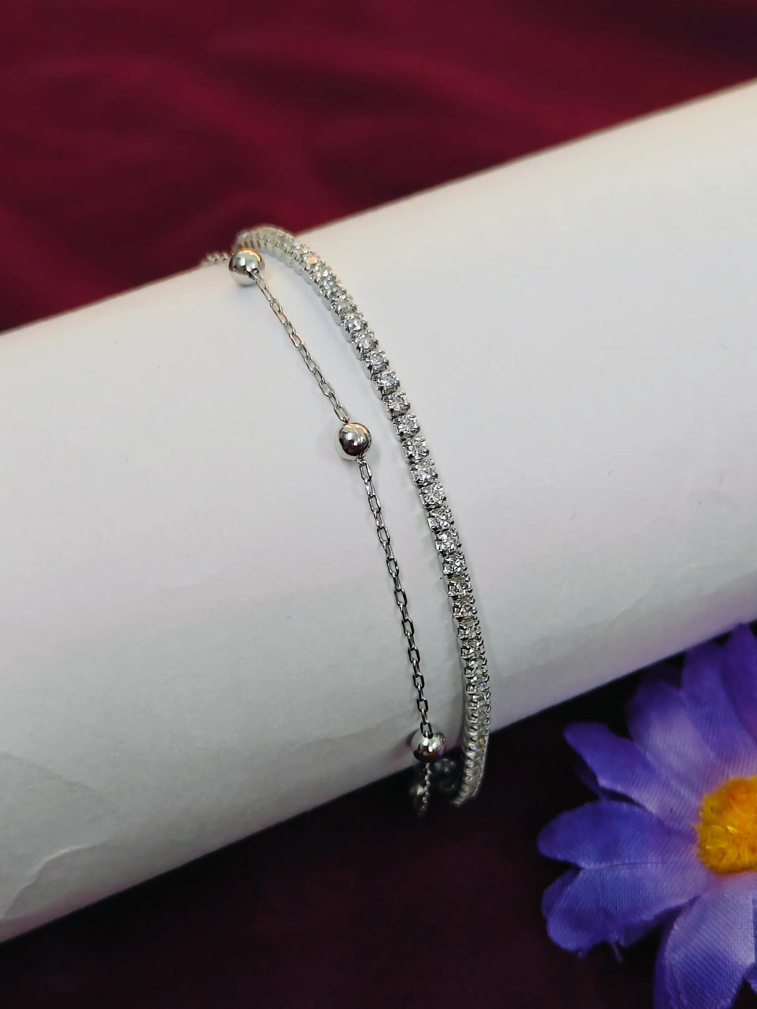 Two silver bracelets on a white surface with a purple flower in the background