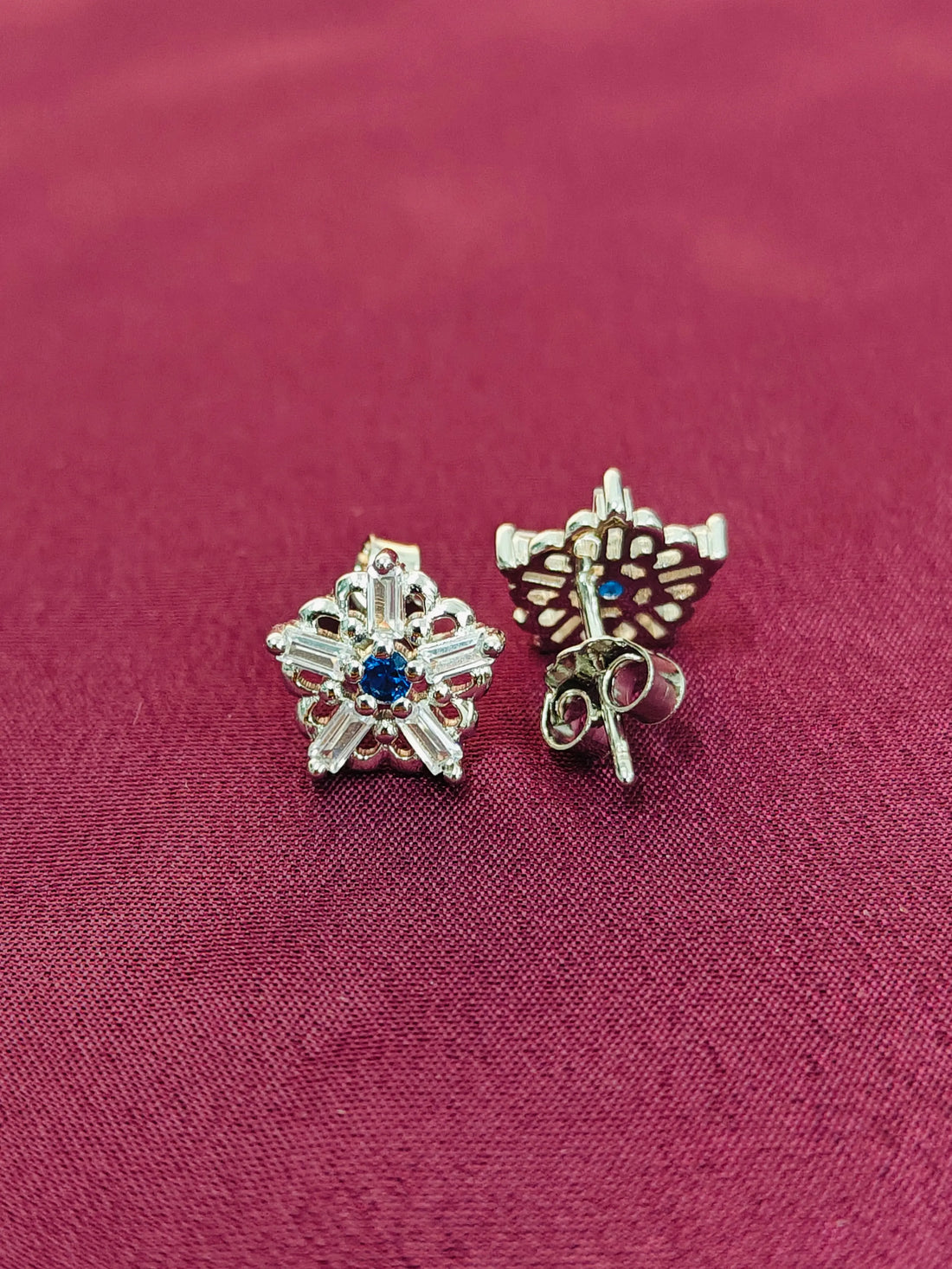 Two silver star-shaped earrings with blue stones on a red fabric background