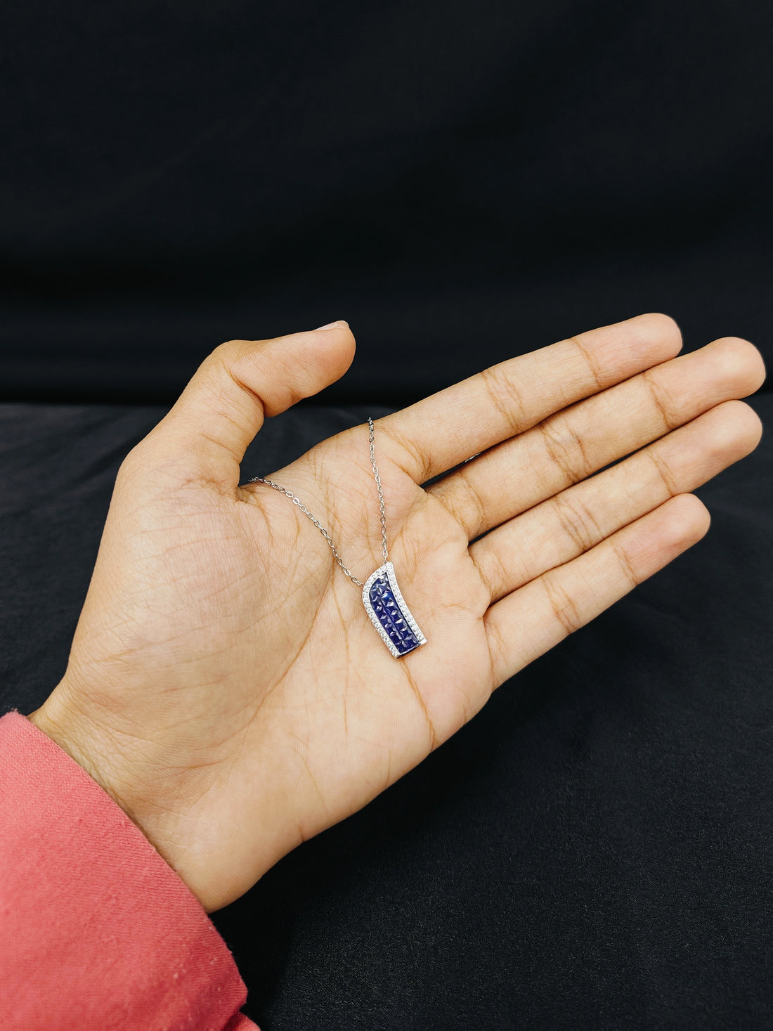 Hand holding a silver necklace with a blue pendant against a black background