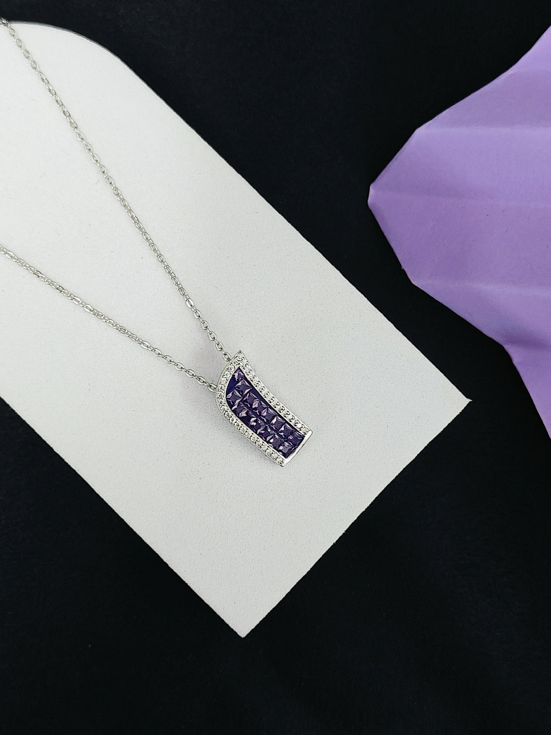 Silver necklace with a purple pendant on a white card against a black background