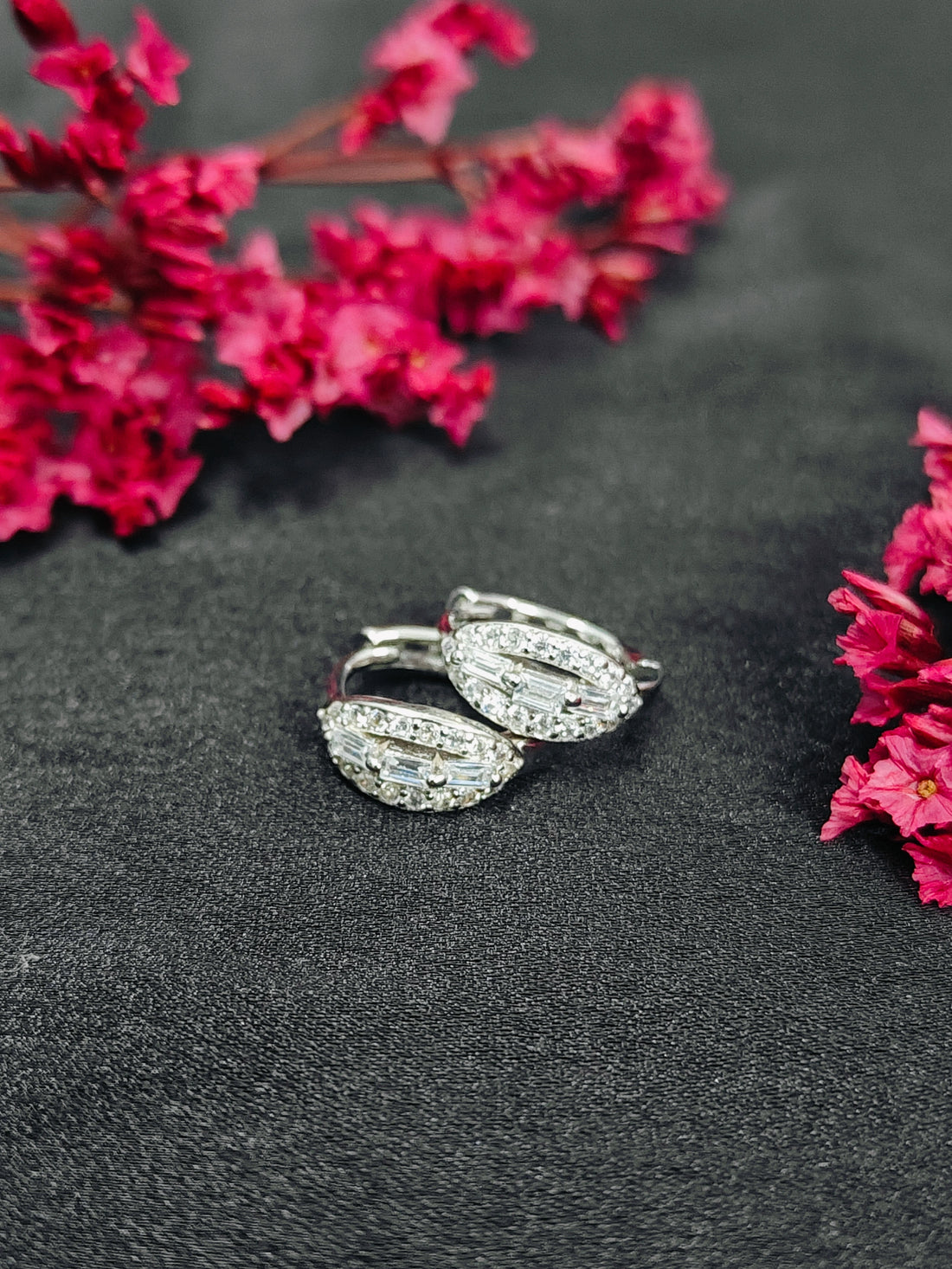 Silver wing-shaped earrings with clear stones on a dark surface with pink flowers.