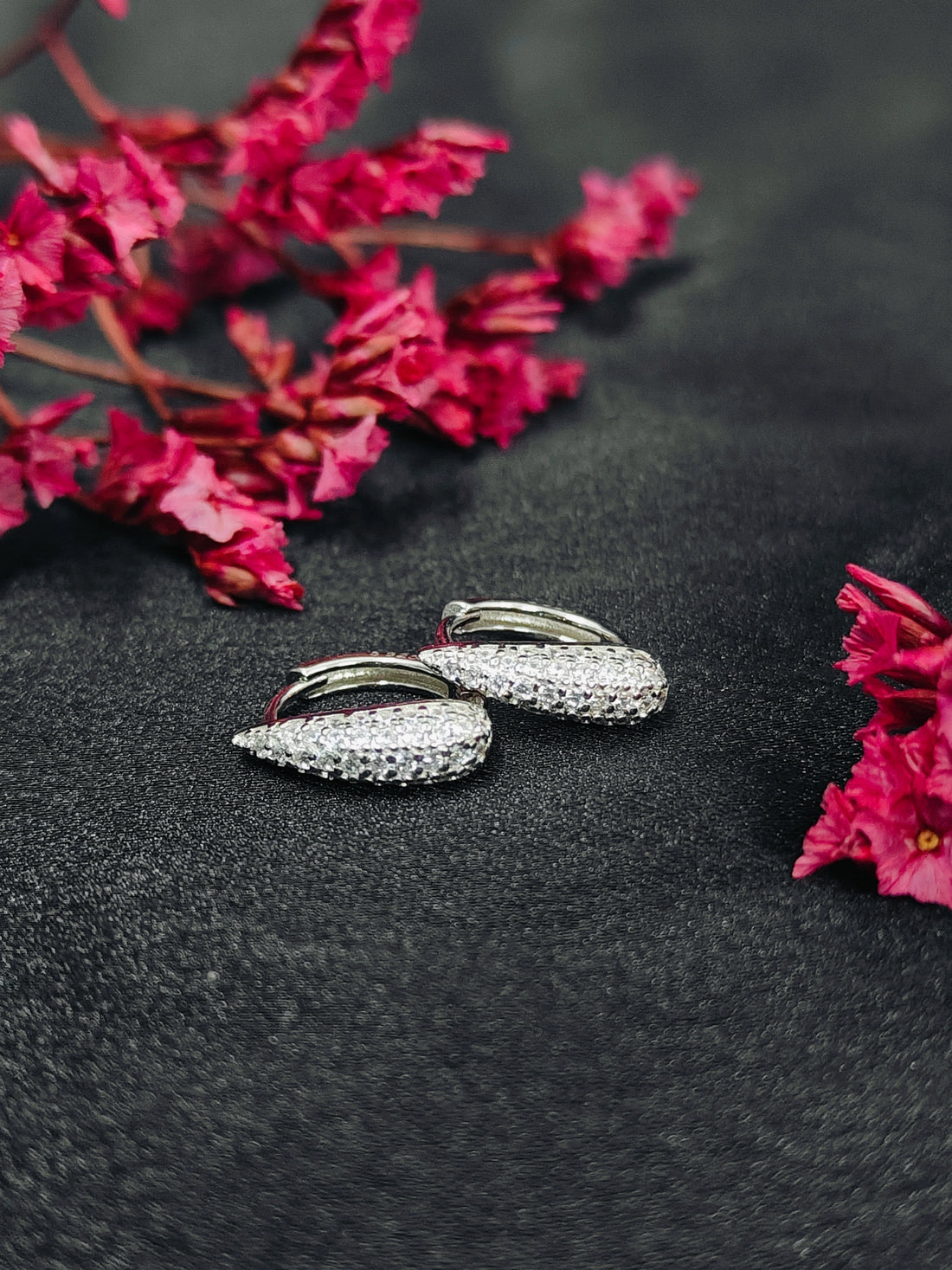 Silver hoop earrings with clear stones on a dark surface with pink flowers.