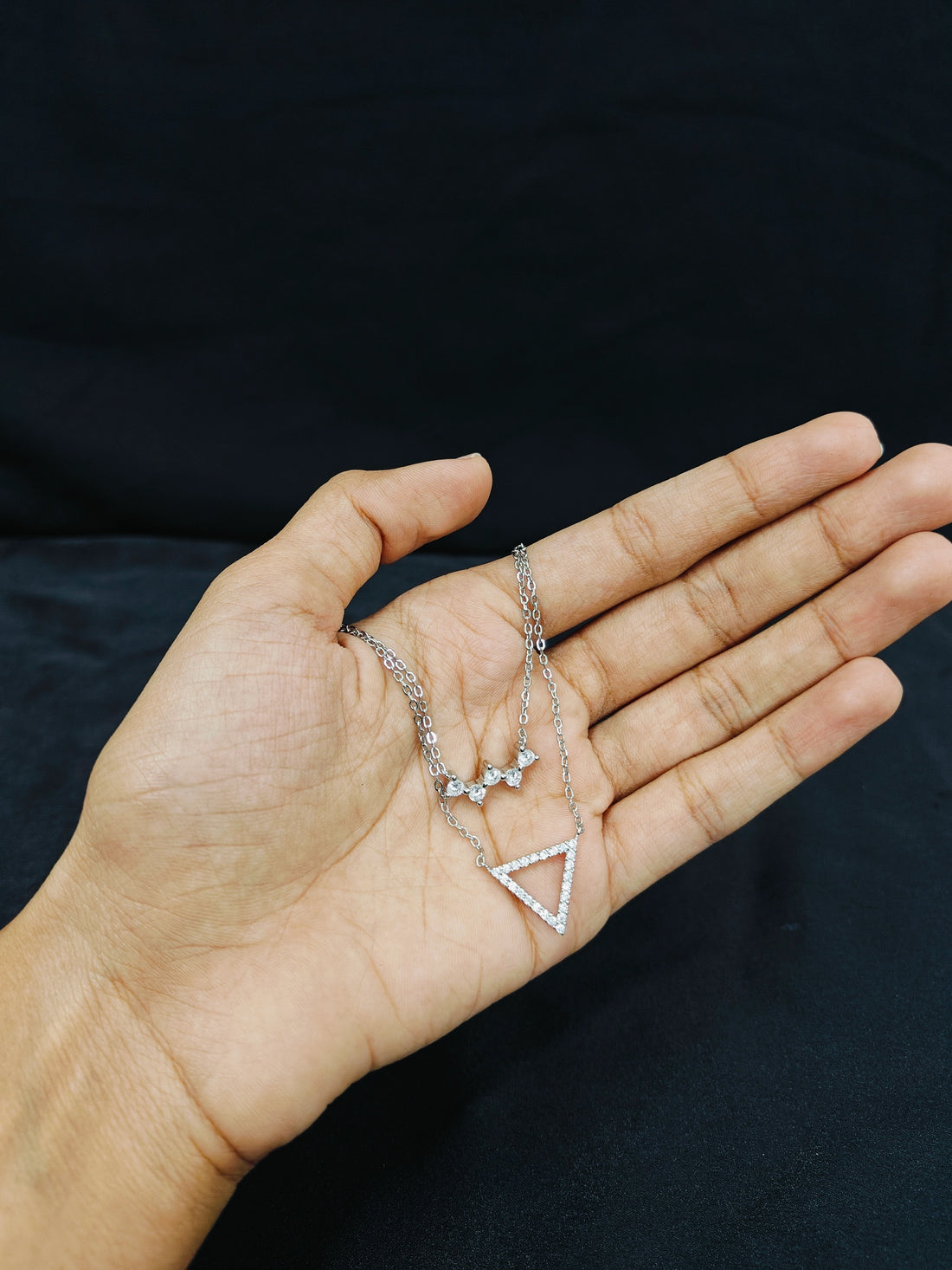 Hand holding a silver necklace with a triangle pendant against a black background