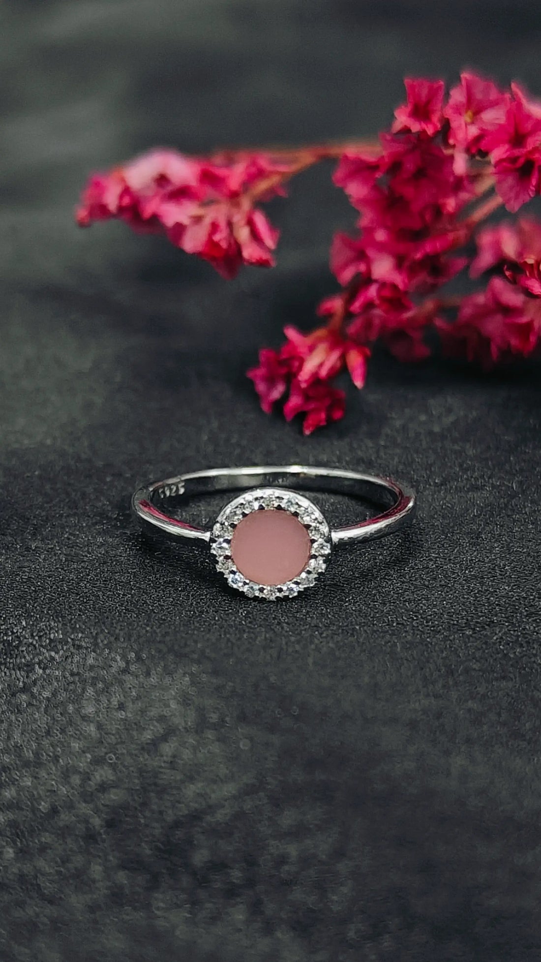 Silver ring with a pink gemstone on a dark surface with red flowers in the background