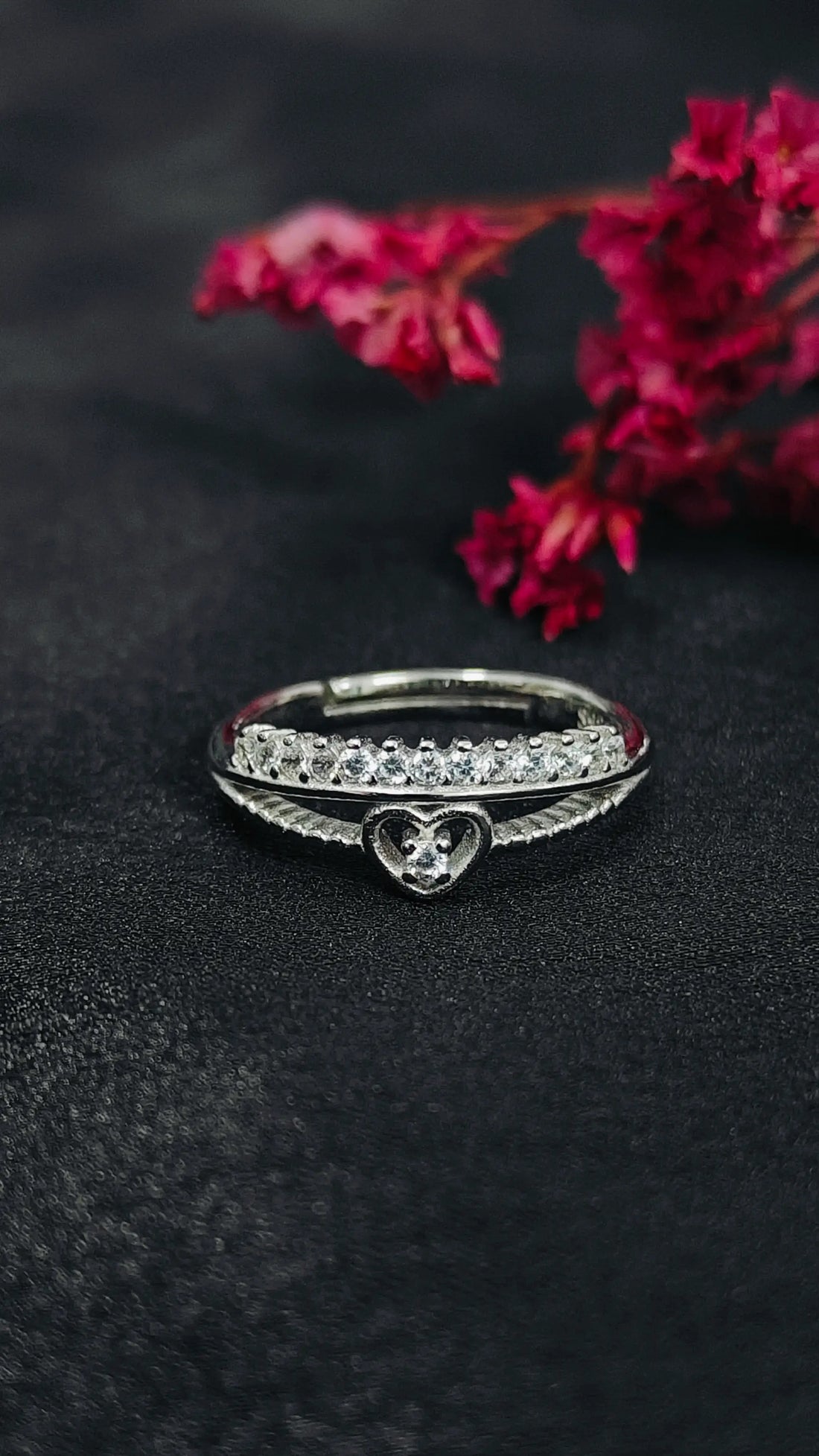 Silver ring with heart design on a black surface with pink flowers in the background