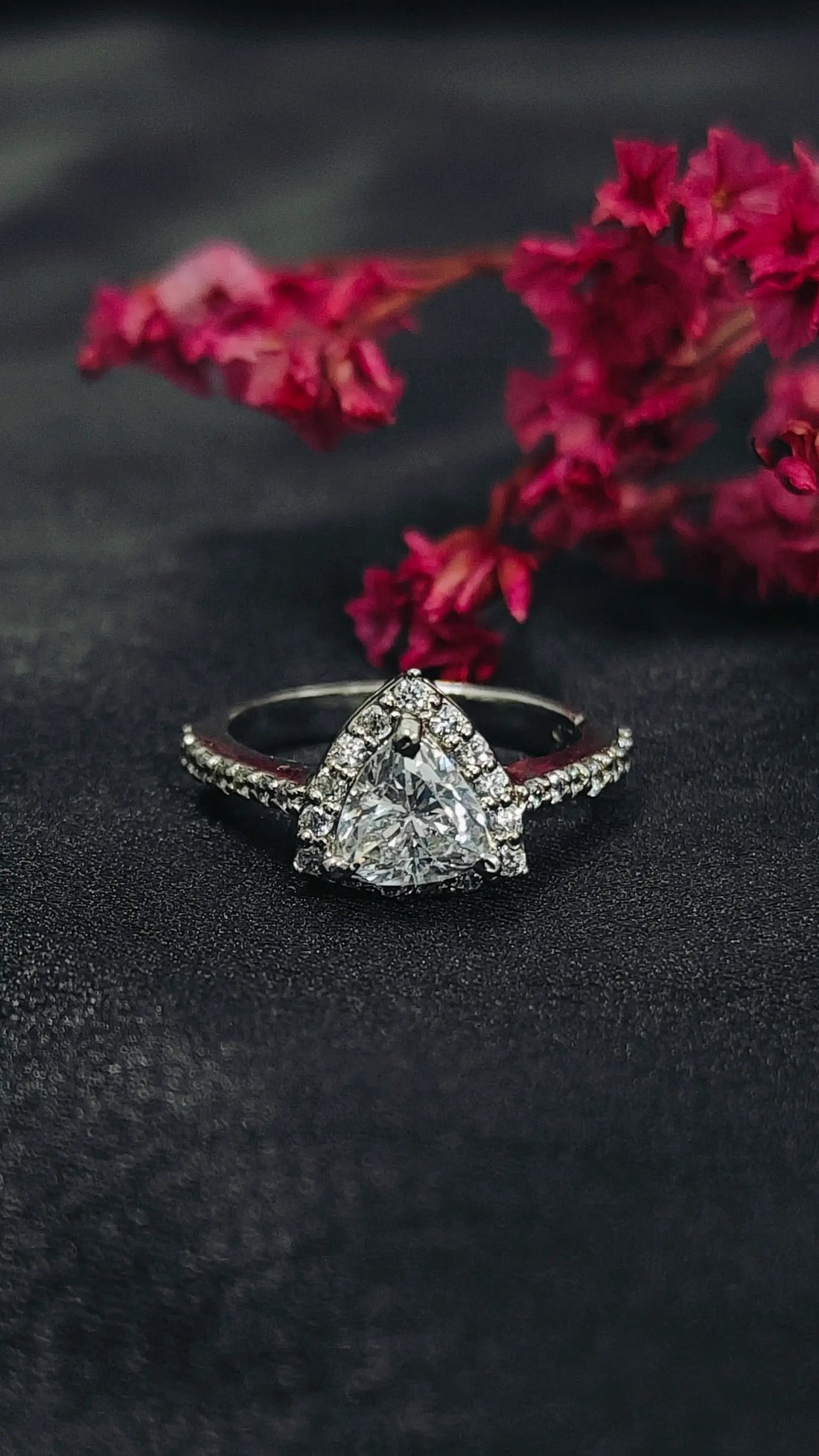 Diamond ring with a triangle-shaped center stone on a dark surface with pink flowers.