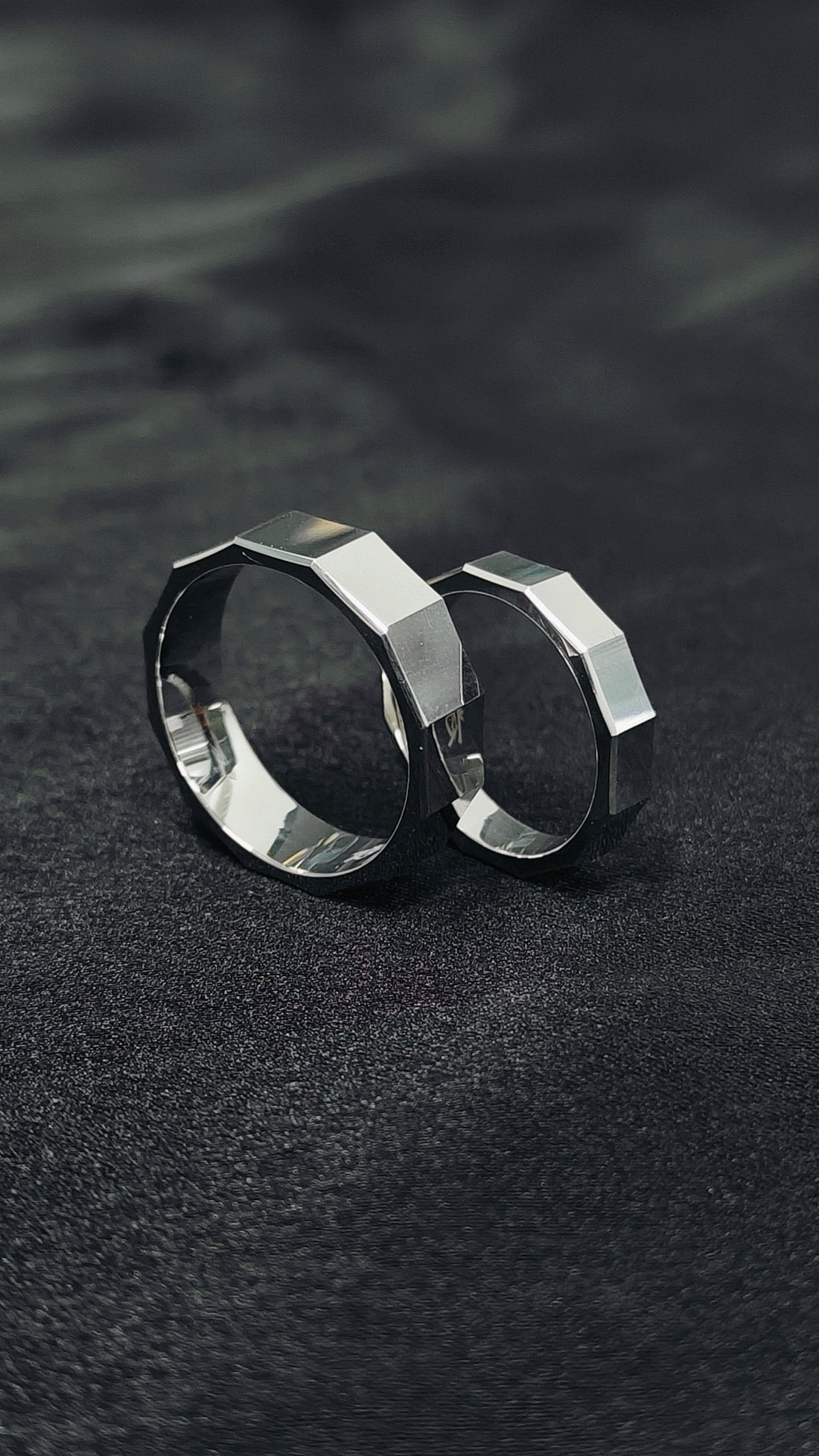 Two silver rings with geometric designs on a dark textured surface