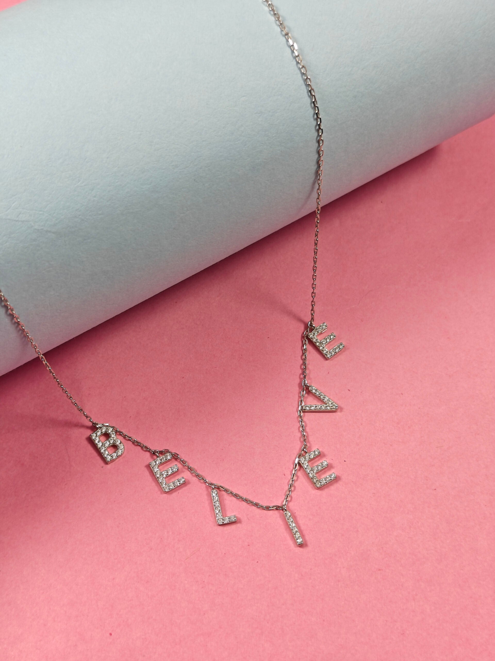 Necklace with 'BELIEVE' pendant on a pink and gray background