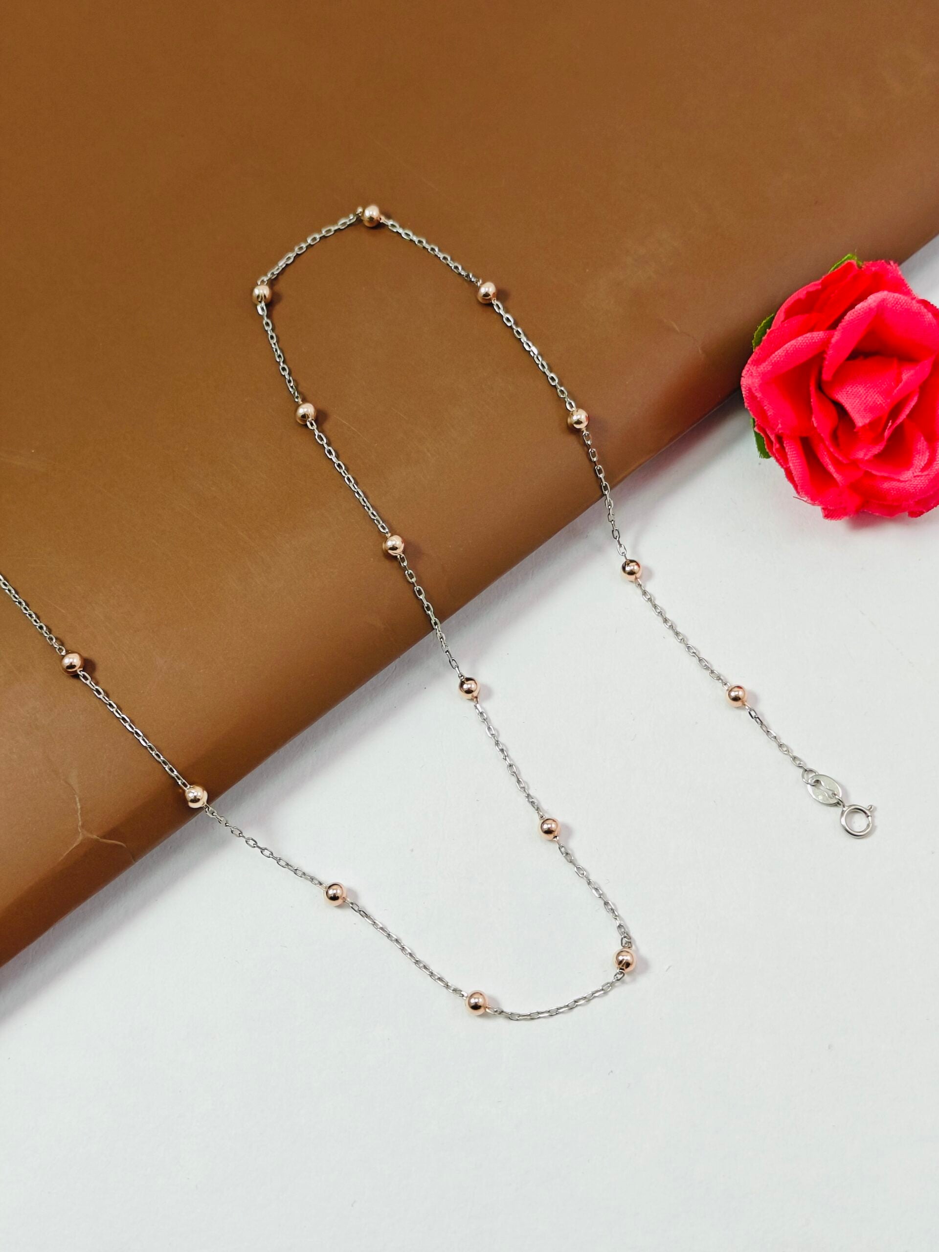 Silver necklace with small beads on a white surface with a pink flower.