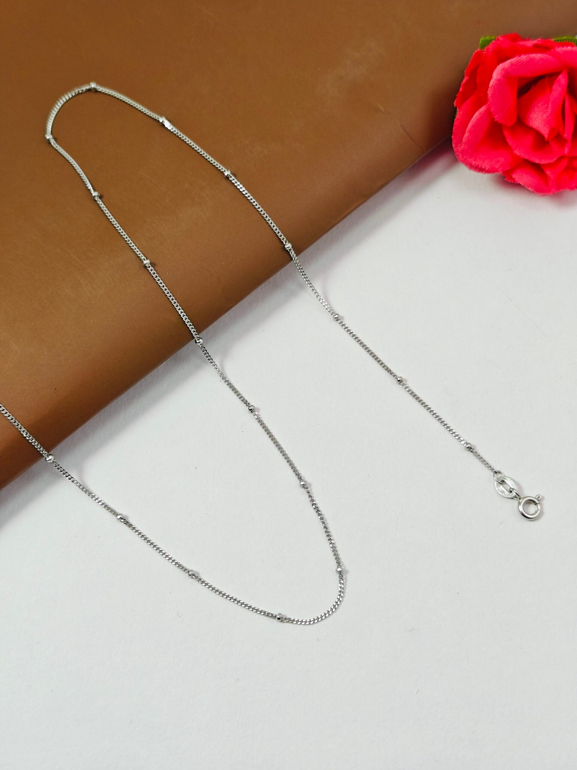 Silver chain necklace on a white surface with a red flower in the corner