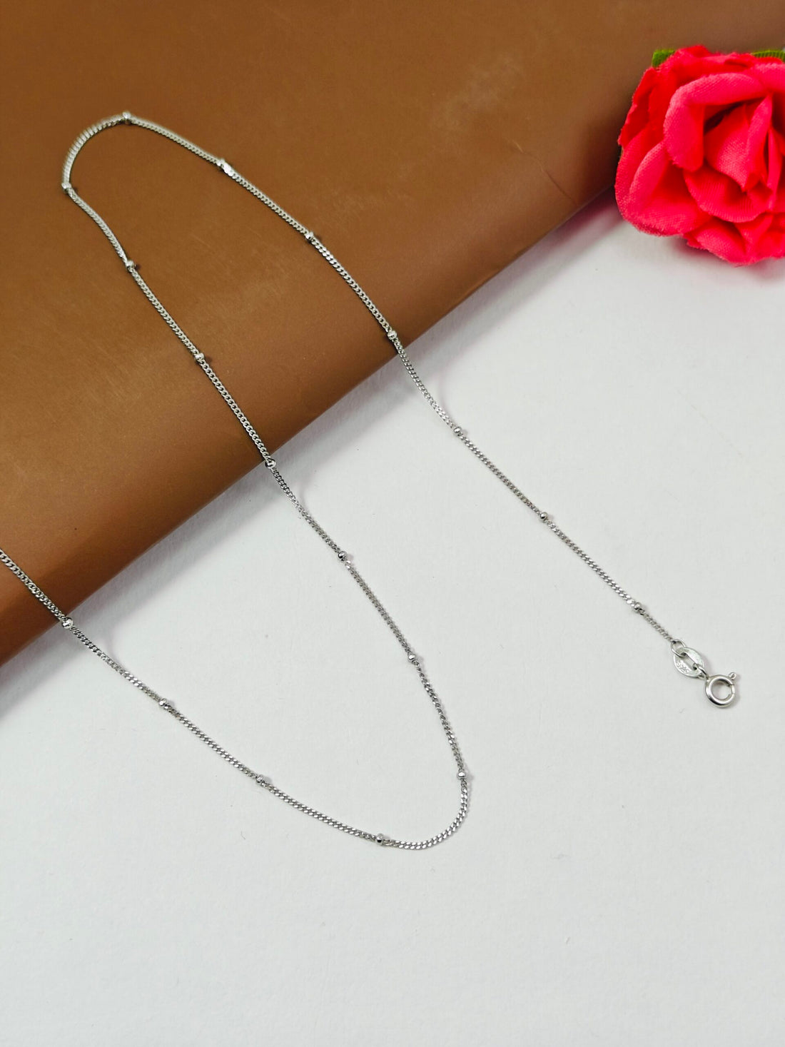 Silver chain necklace on a white surface with a red flower in the corner