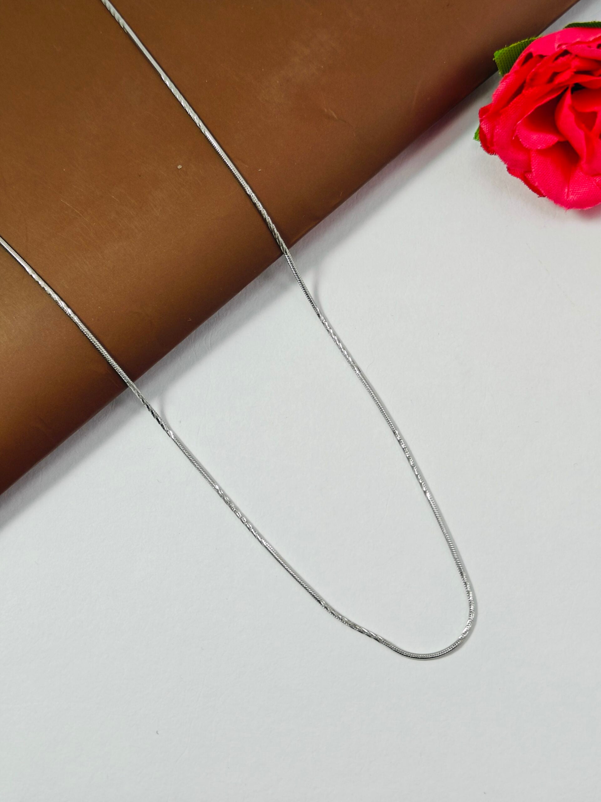 Silver chain necklace on a white surface with a red flower in the corner