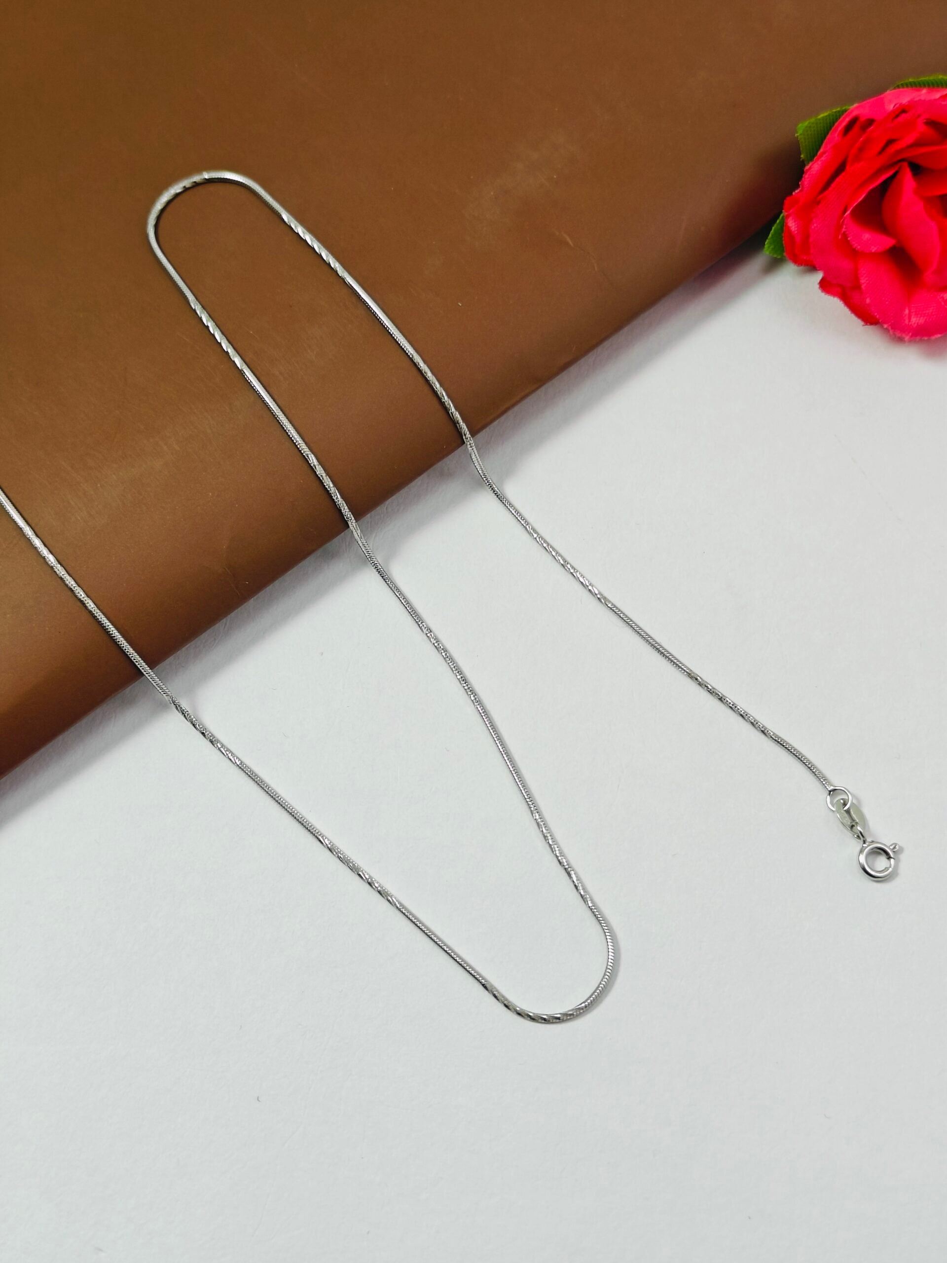 Silver chain necklace on a white surface with a red flower in the corner
