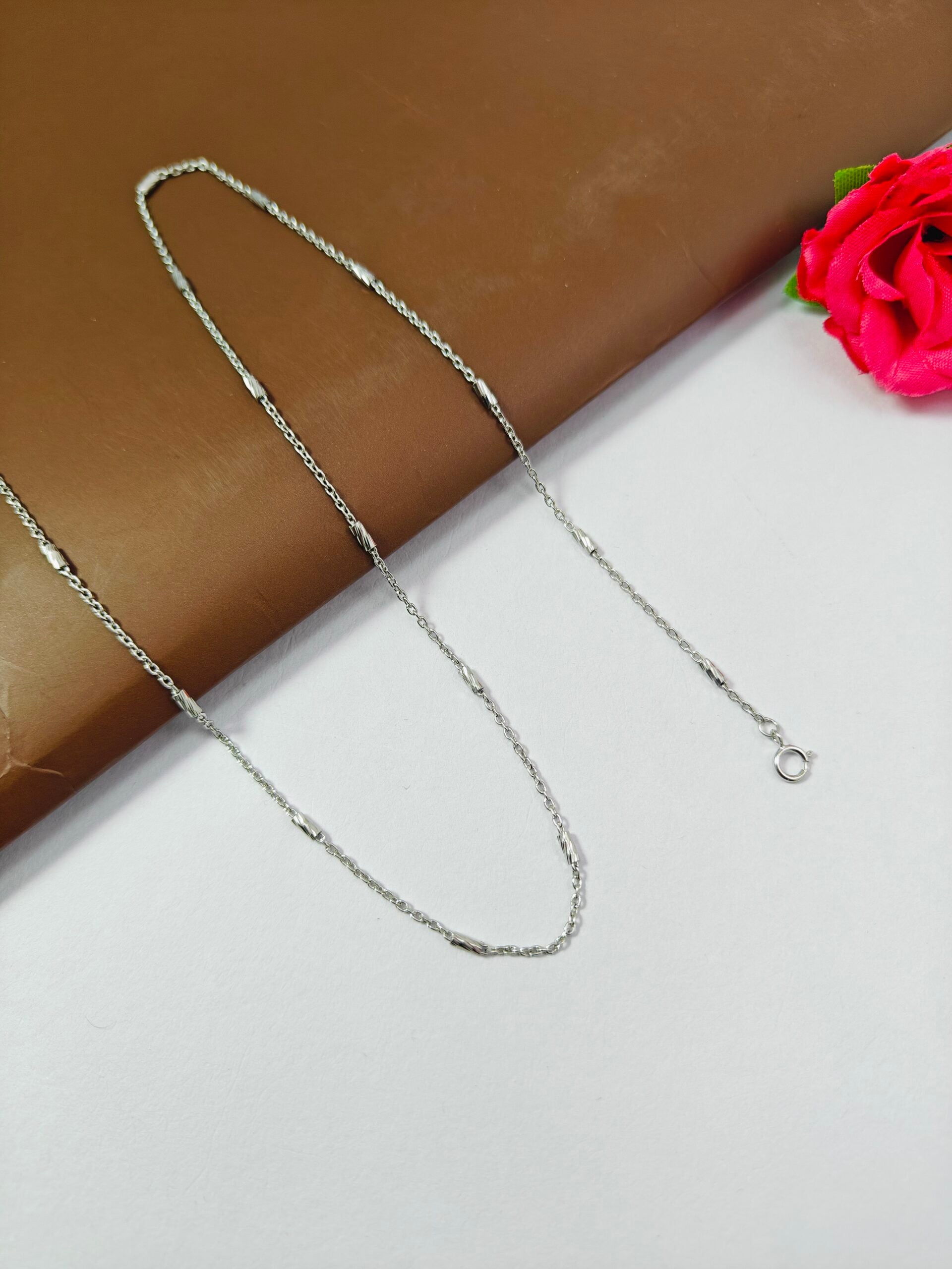 Silver chain necklace on a white surface with a pink flower in the corner