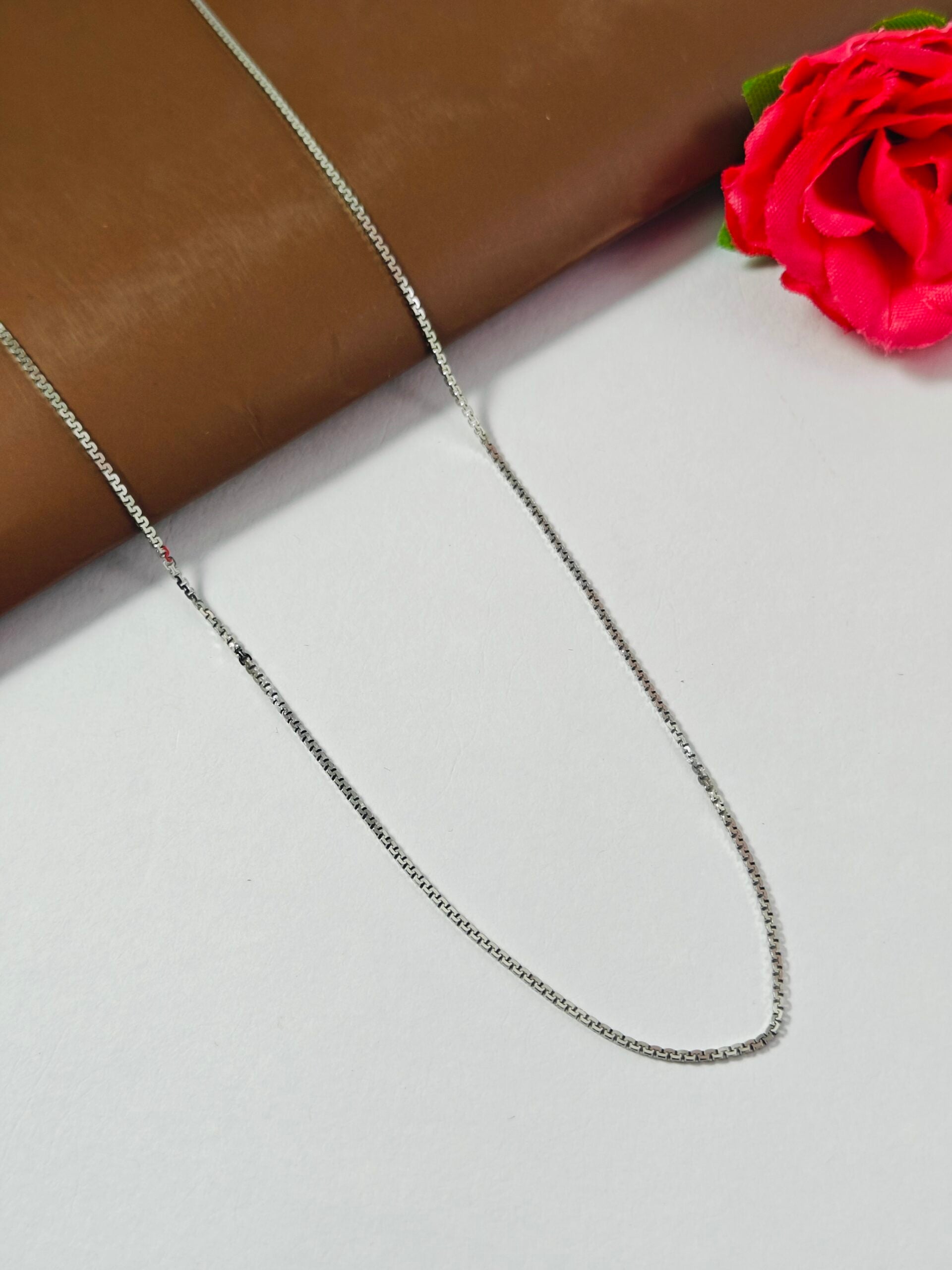 Silver chain necklace on a light gray surface with a brown fabric and red flower in the background