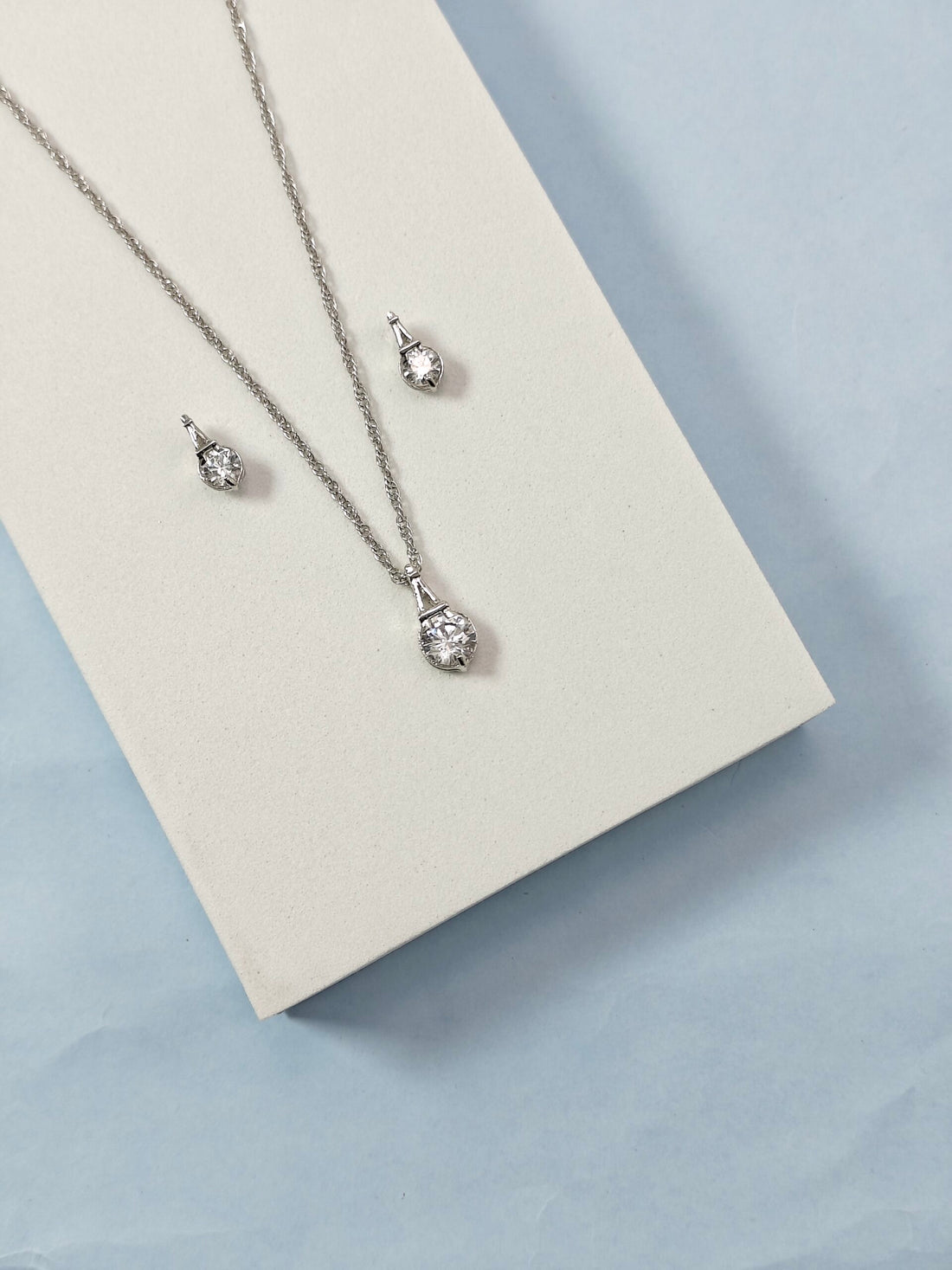 Silver necklace and earrings set on a beige card with a light blue background