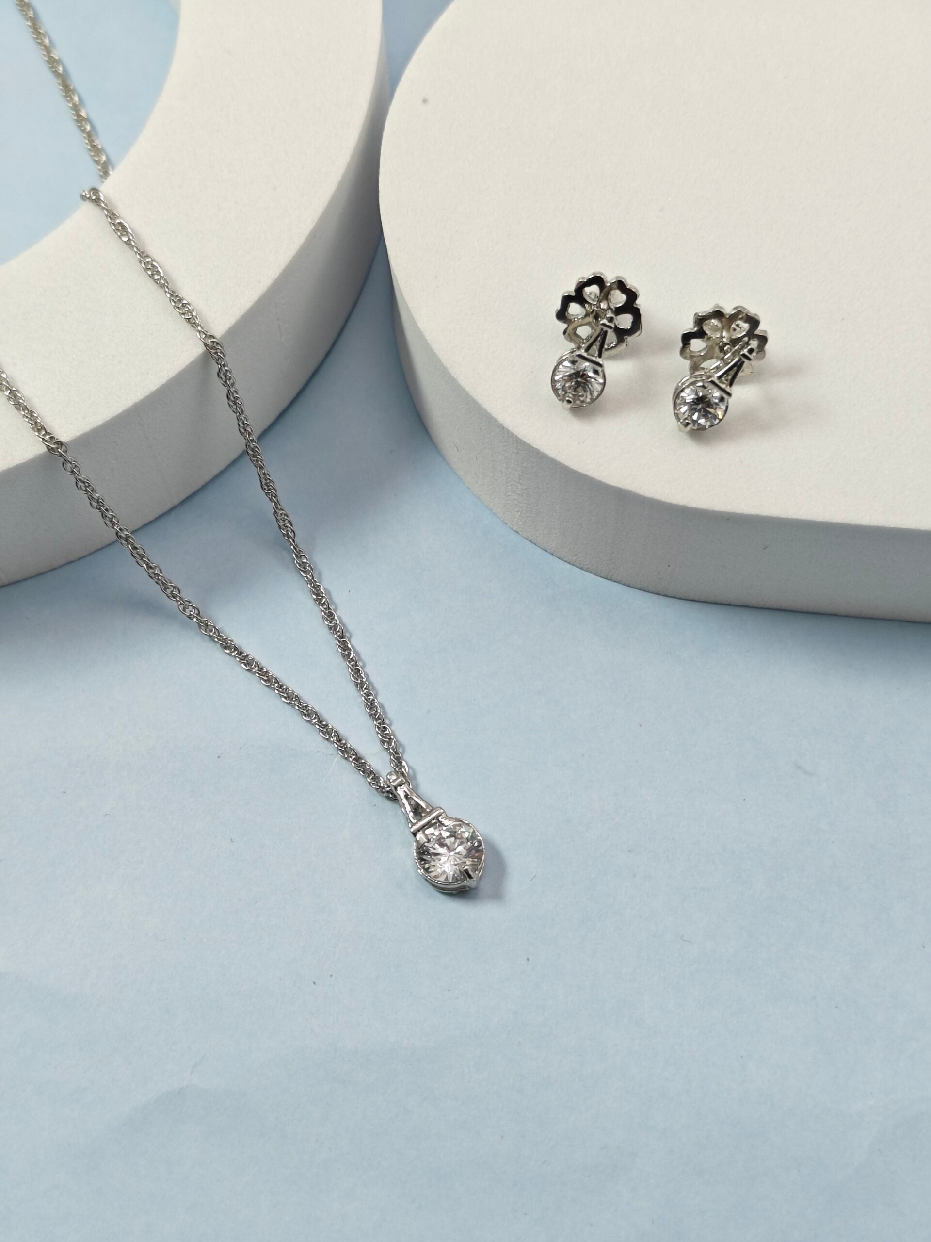 Silver necklace and earrings on a light blue surface with white circular stands.