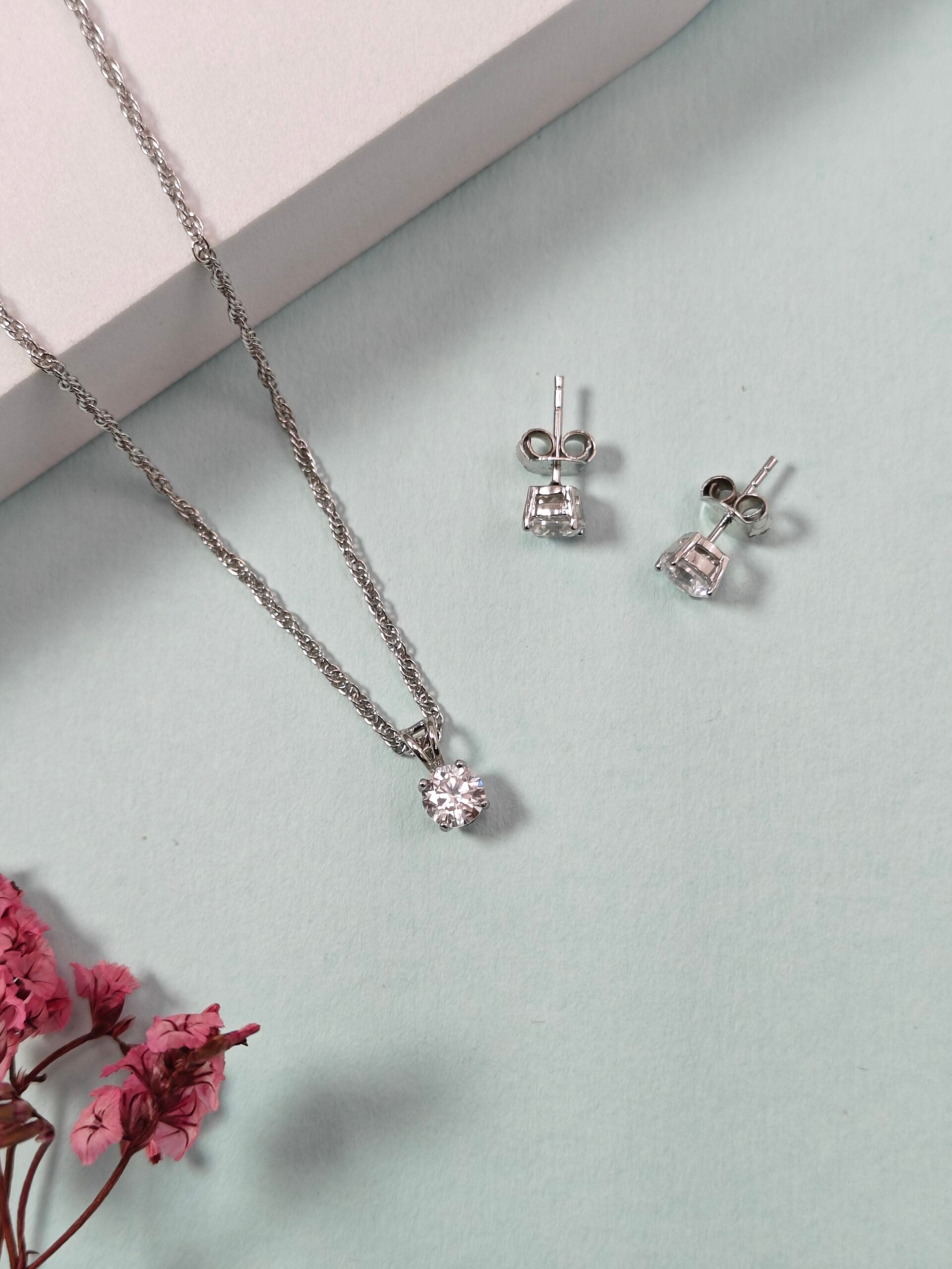 Silver necklace and earrings on a light gray surface with pink flowers.