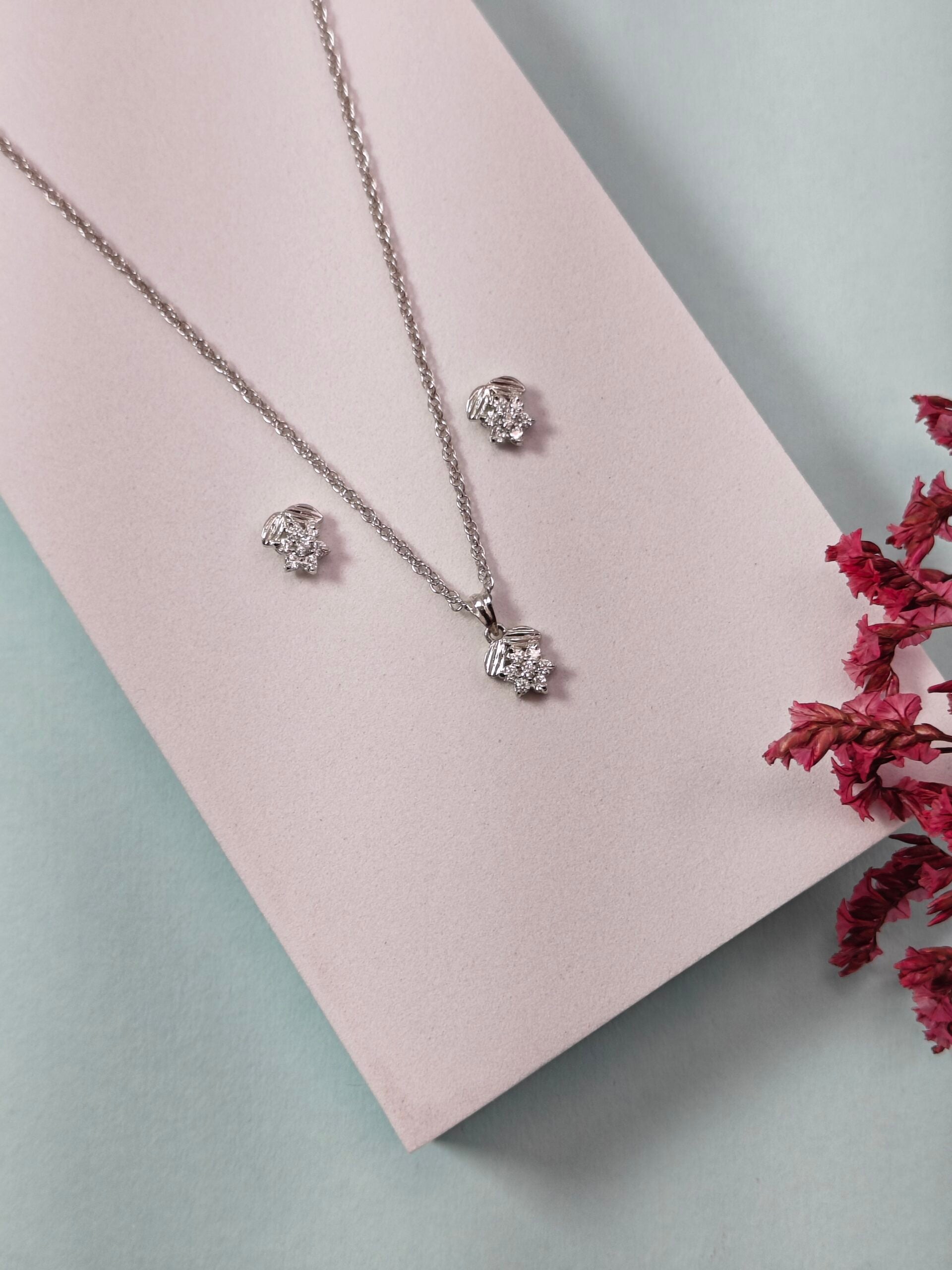 Silver jewelry set with necklace and earrings on a pink cushion, surrounded by red flowers.