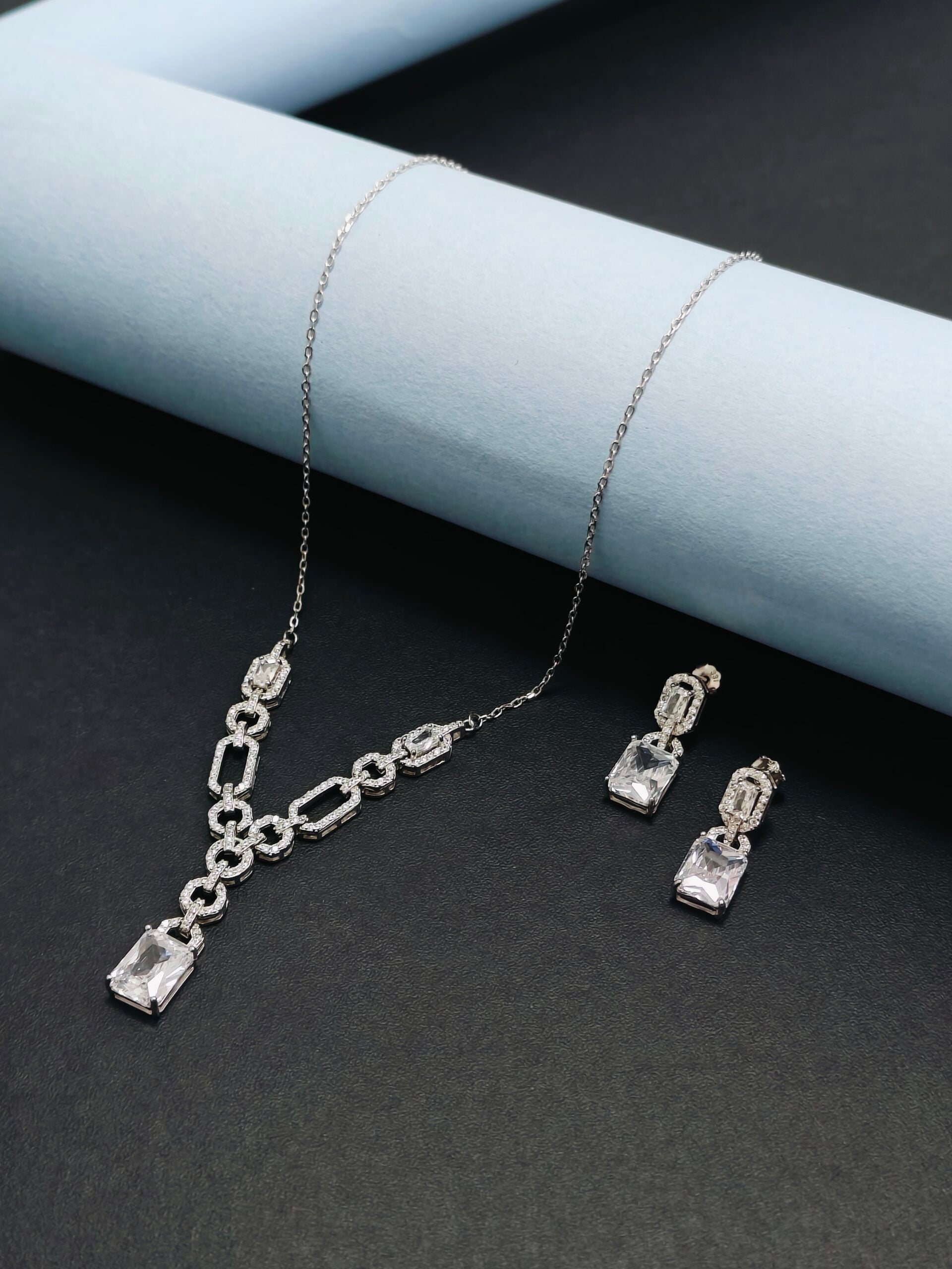 Silver necklace and earrings set on a dark surface with light blue cylindrical object.