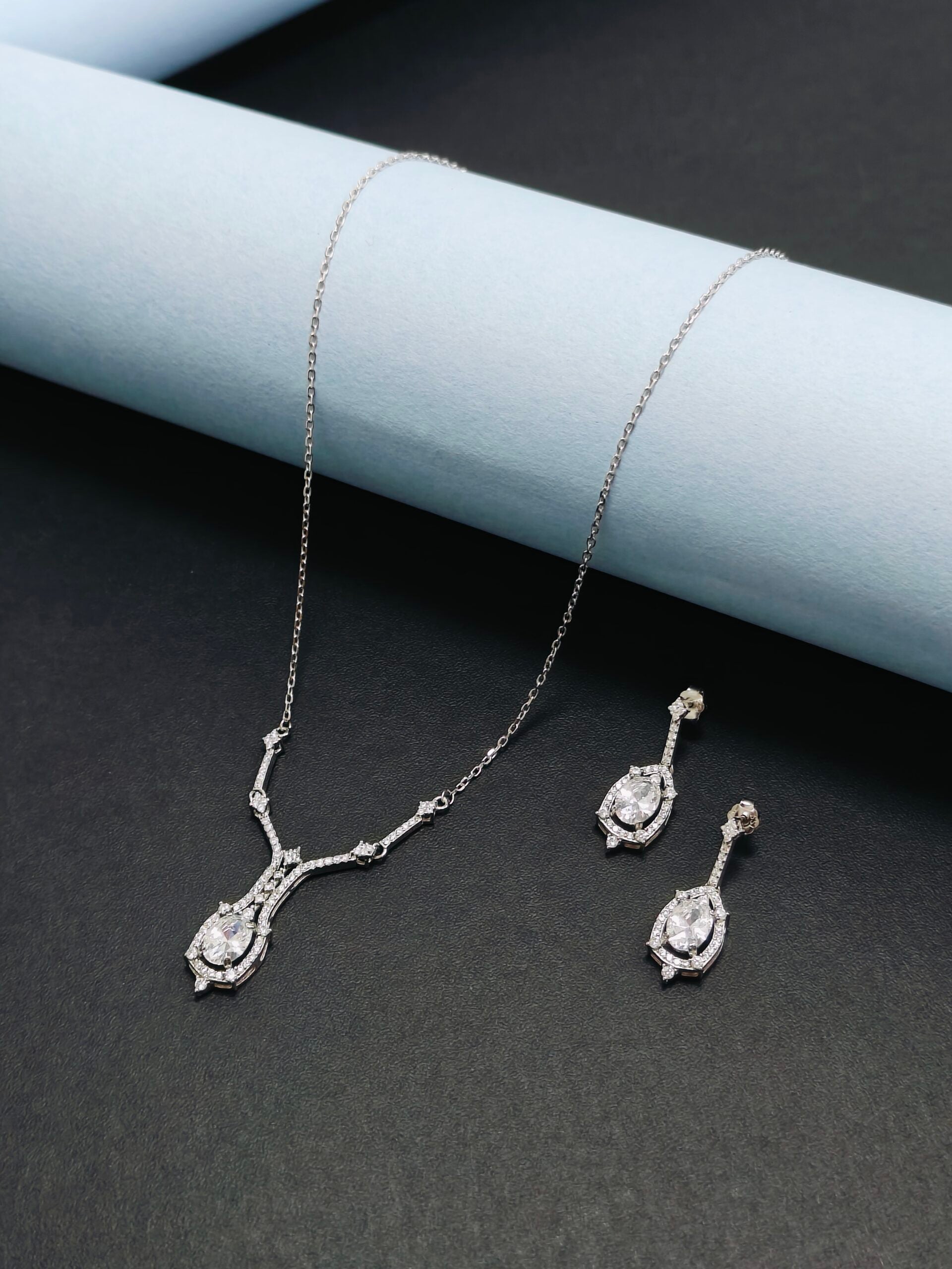 Silver necklace and earrings set on a dark surface with light blue cylindrical object.