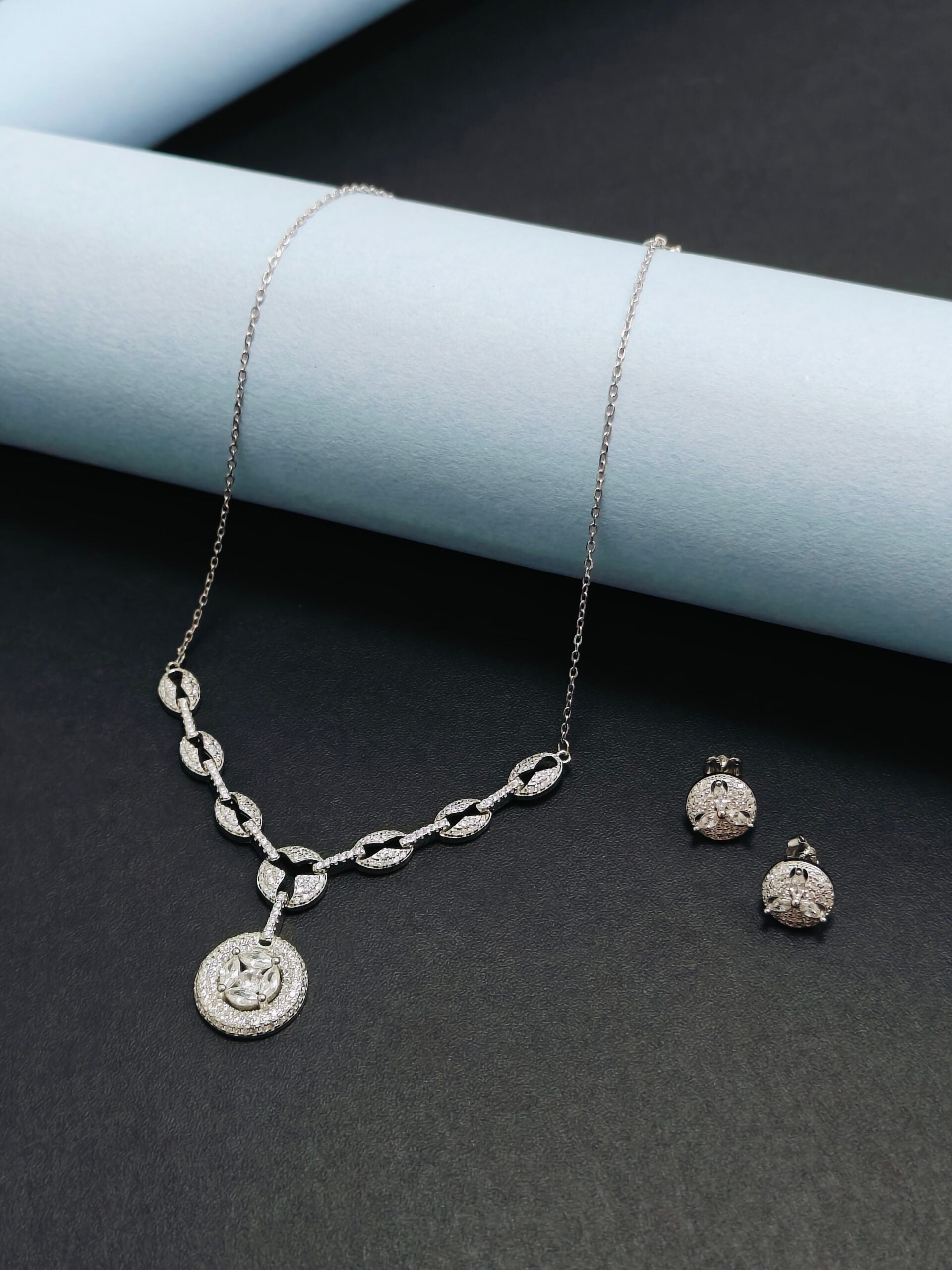 Silver necklace and earrings on a dark surface with a light blue cylindrical object.