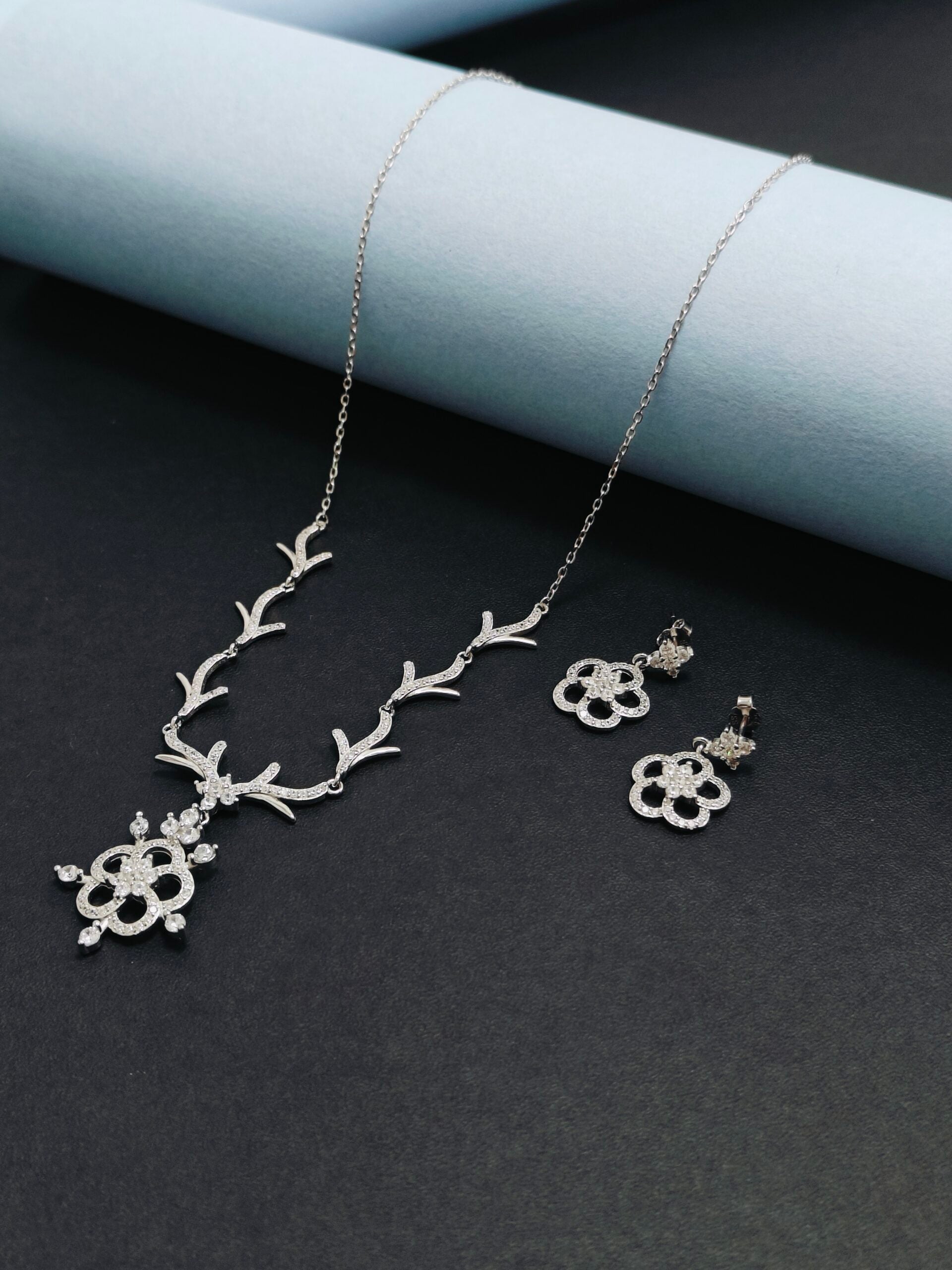 Silver necklace and earrings on a dark surface with a light blue cylindrical object.