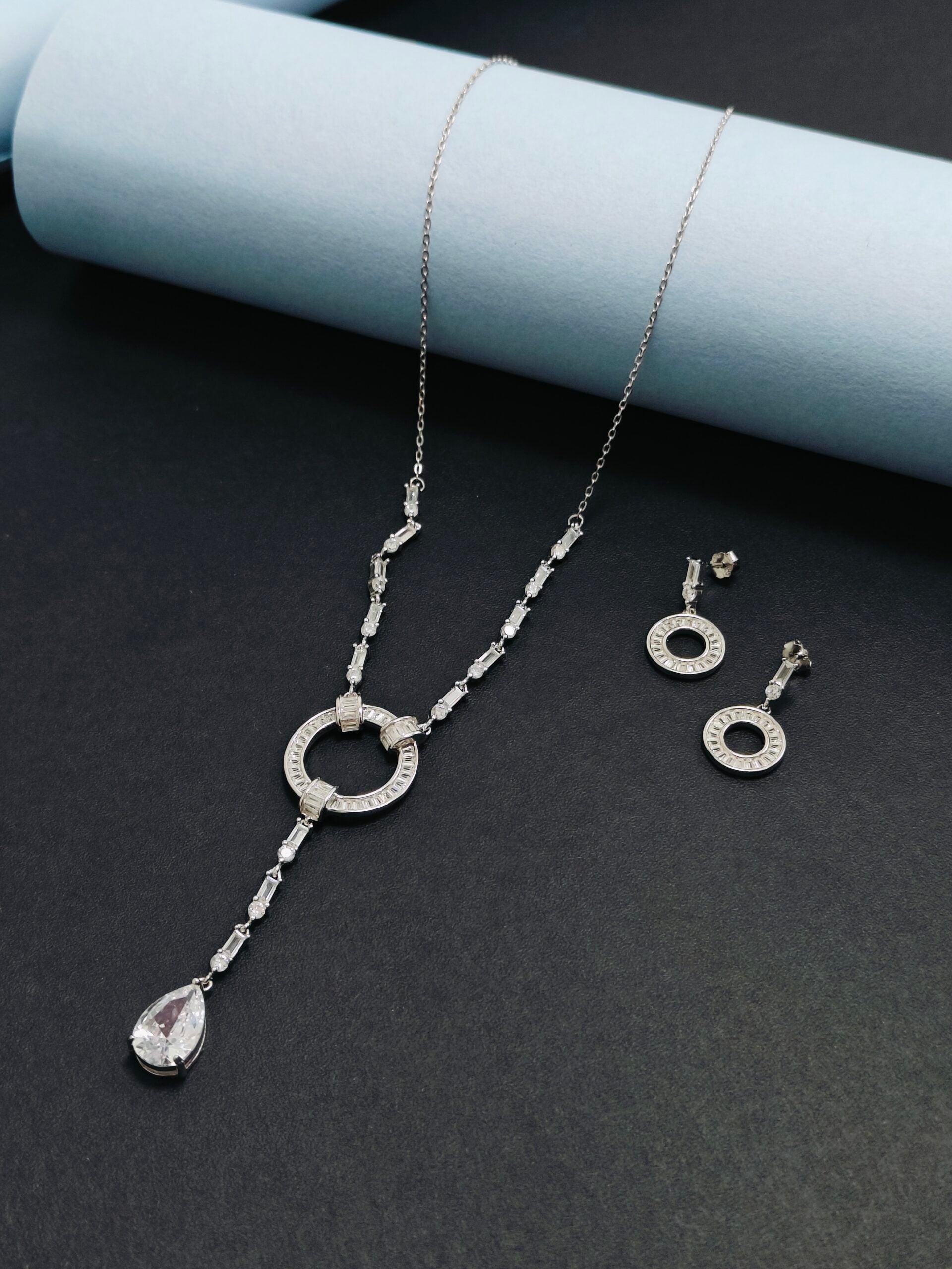Silver necklace and earrings set on a dark surface with a light blue cylindrical object.