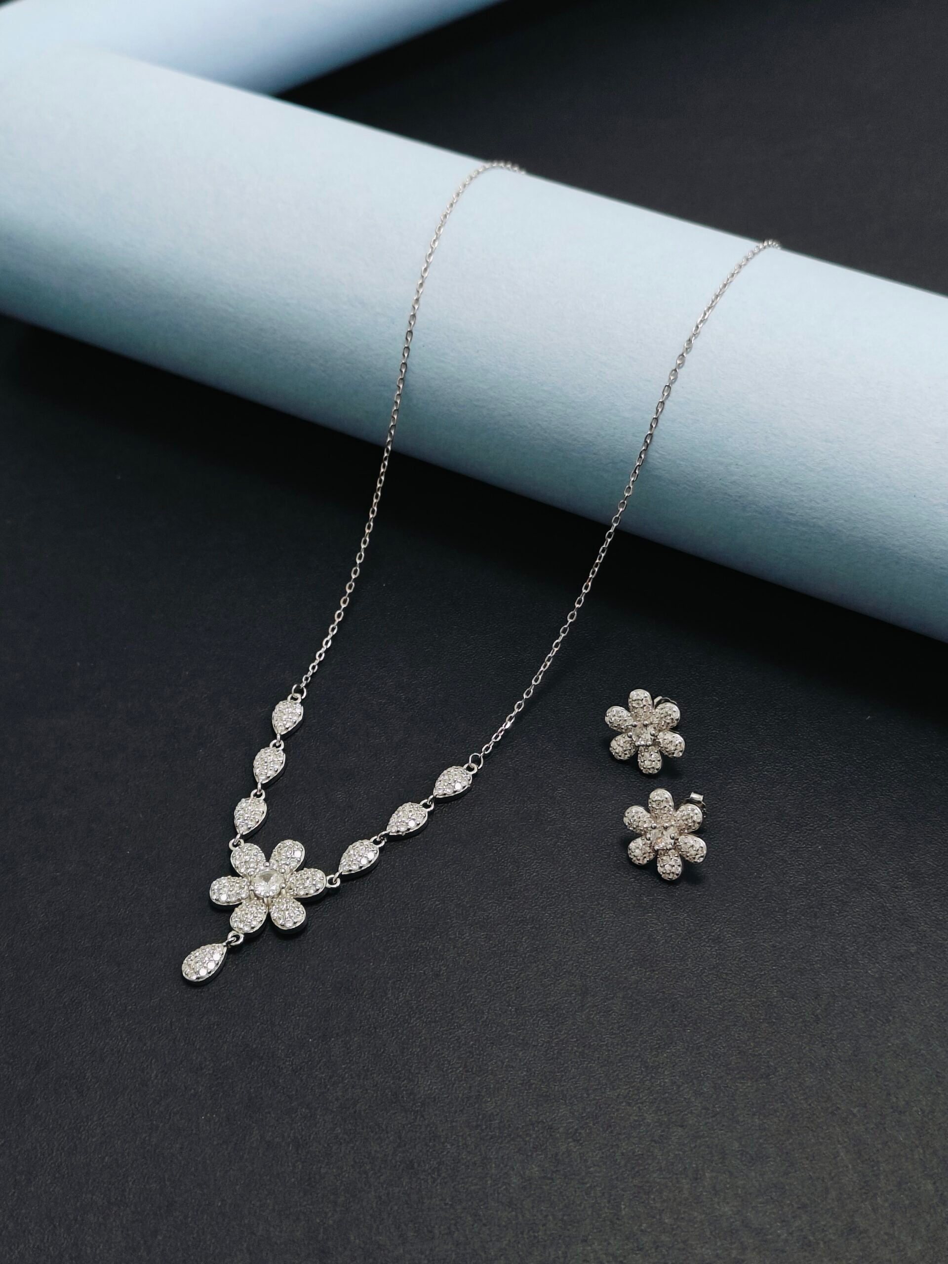 Silver floral necklace and earrings on a dark surface with a light blue cylindrical object.