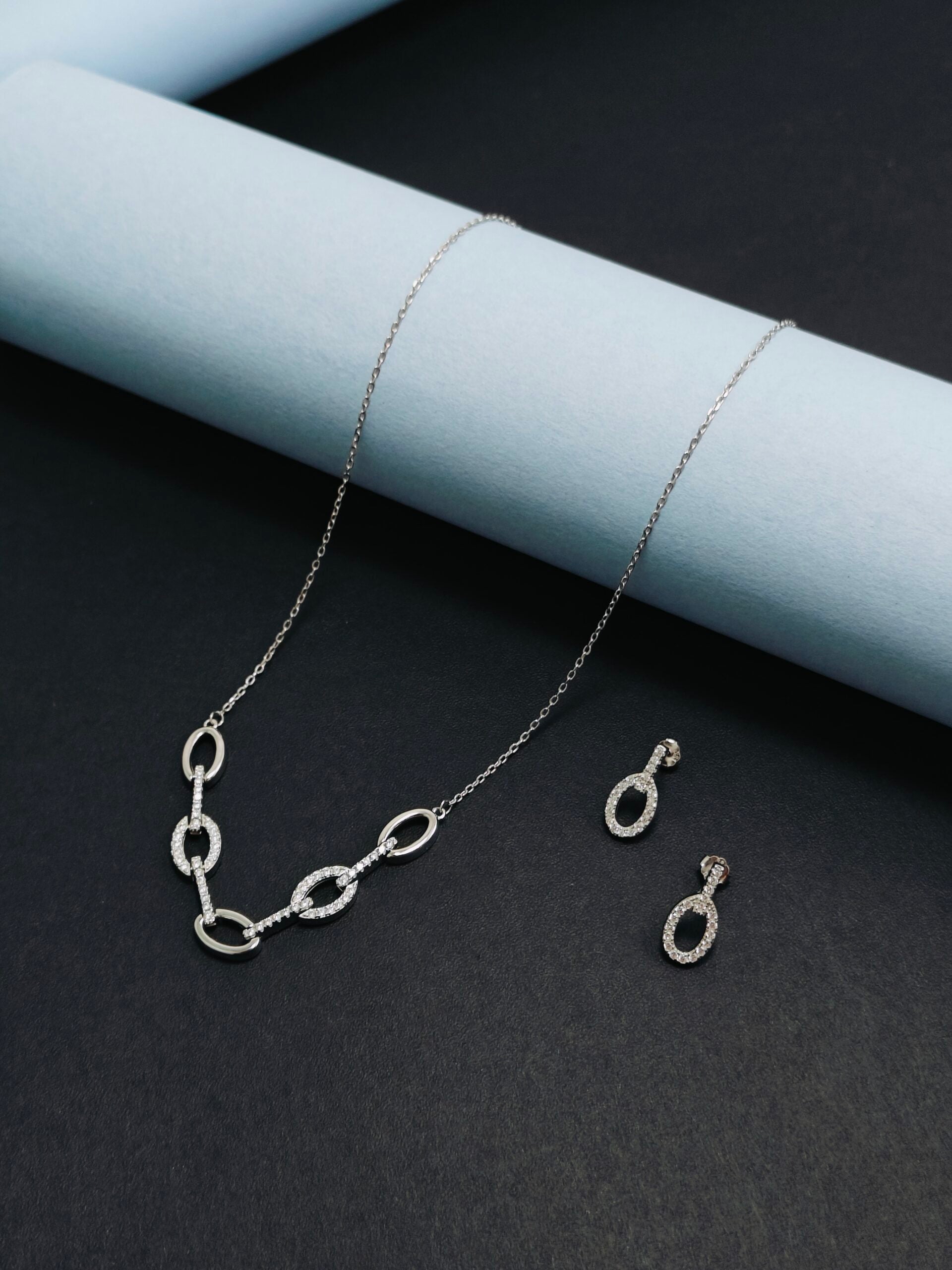 Silver necklace and earrings on a dark surface with light blue cylindrical object