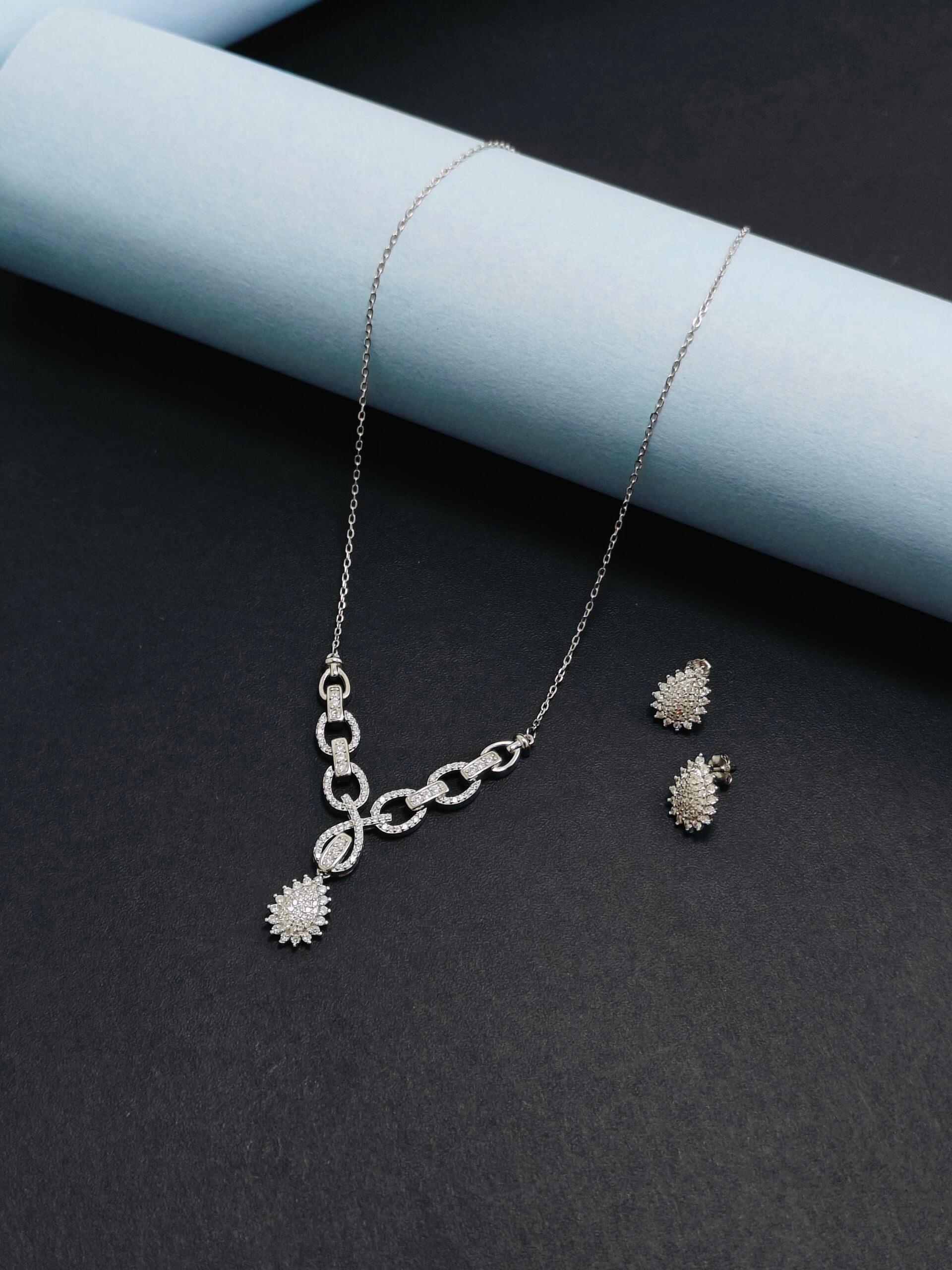 Silver necklace and earrings on a dark surface with a light blue cylindrical object.