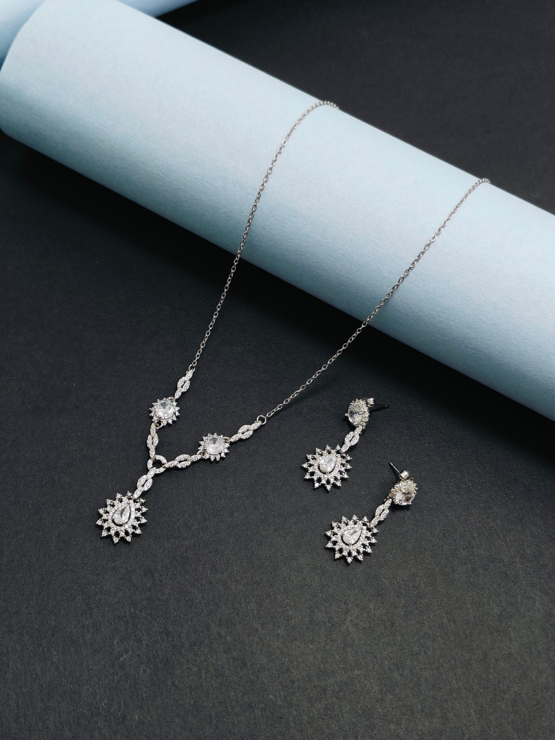 Silver sun-shaped necklace and earrings on a dark surface with a light blue cylindrical object.