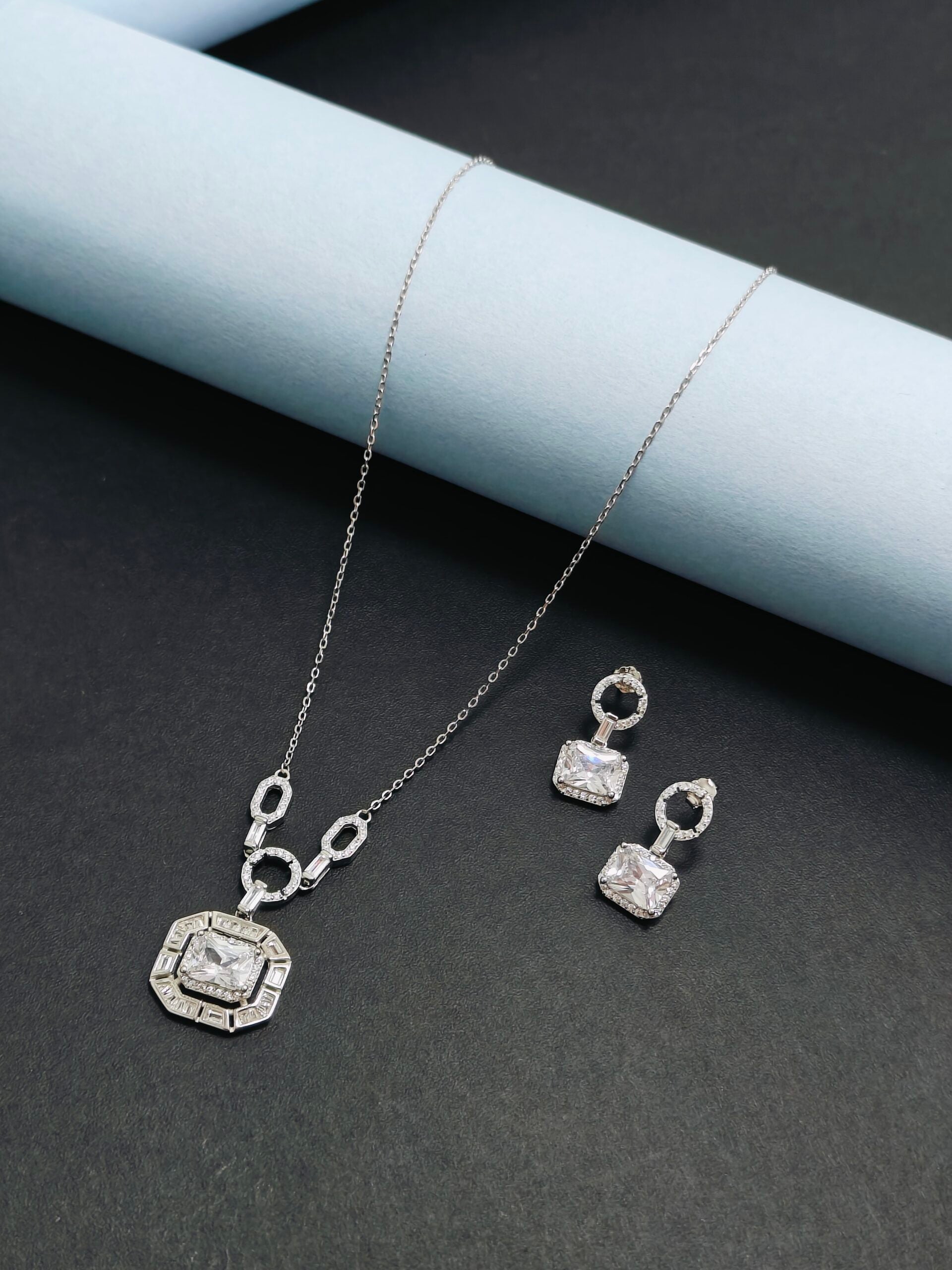 Silver necklace and earrings set on a dark surface with a light blue cylindrical object.