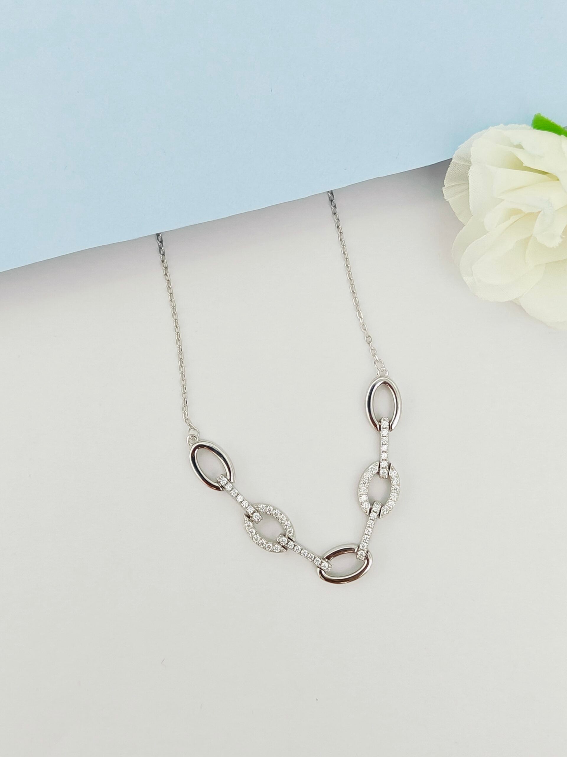 Silver necklace with oval pendants on a light gray surface with a white flower.