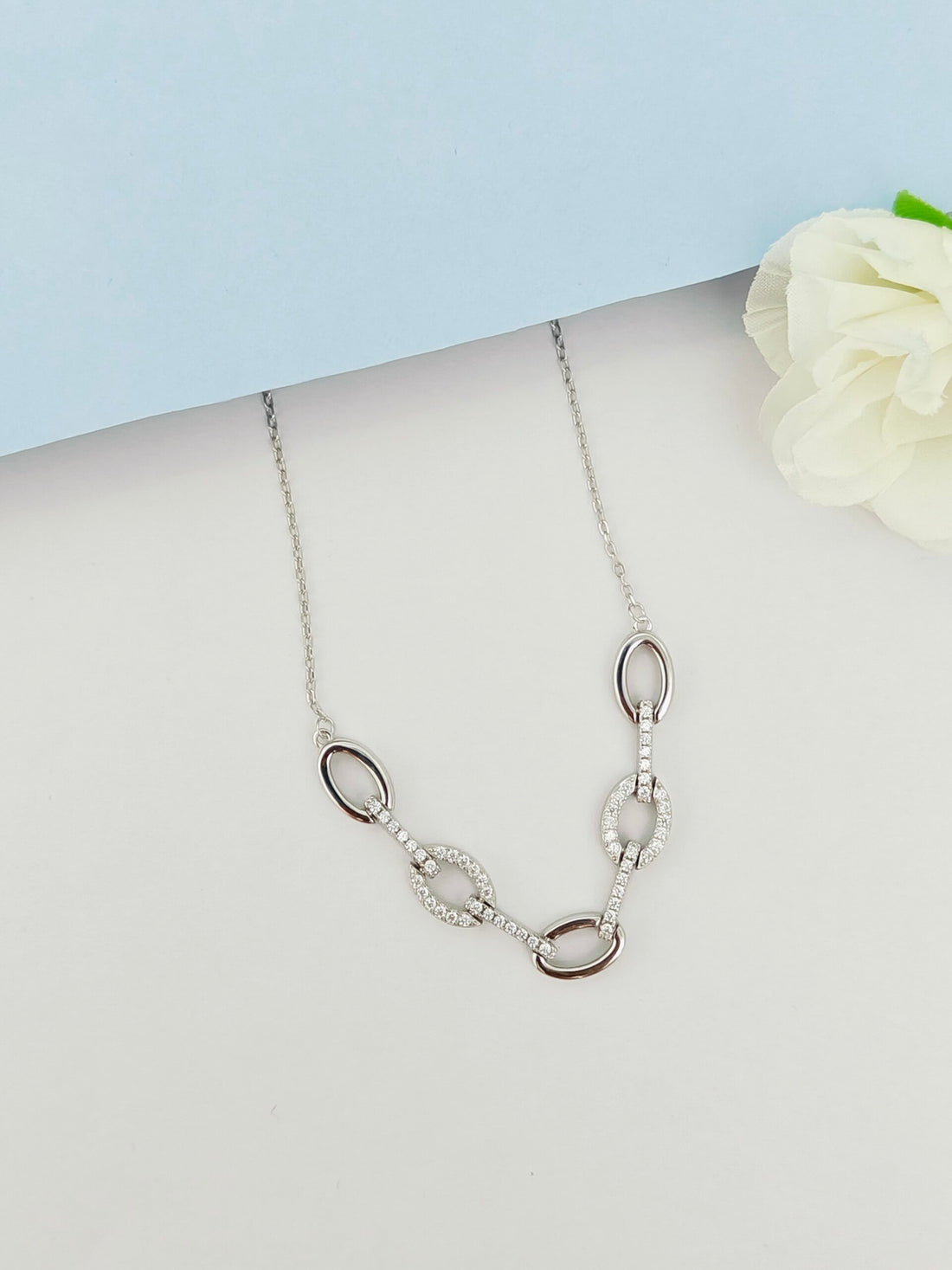 Silver necklace with oval pendants on a light gray surface with a white flower.