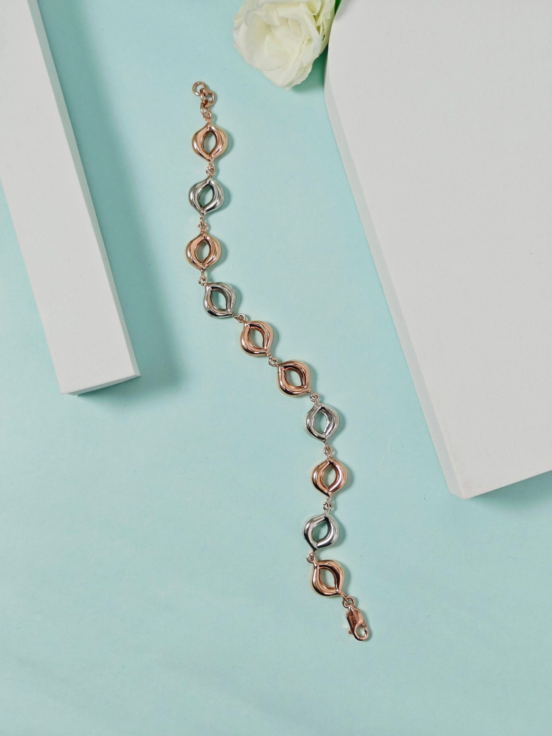 Rose gold bracelet on a light blue surface with a white flower and box in the background