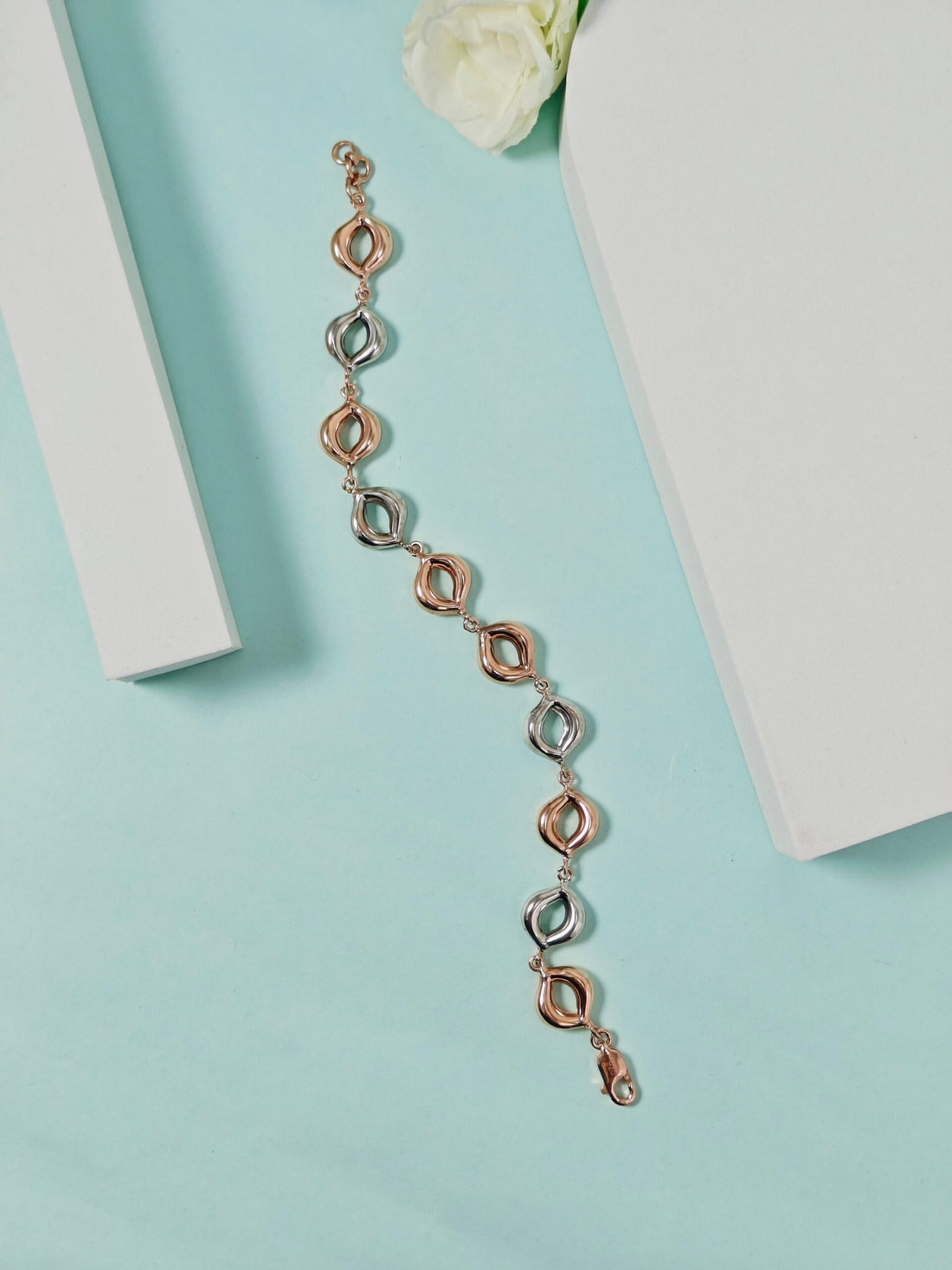 Rose gold bracelet on a light blue surface with a white flower and box in the background
