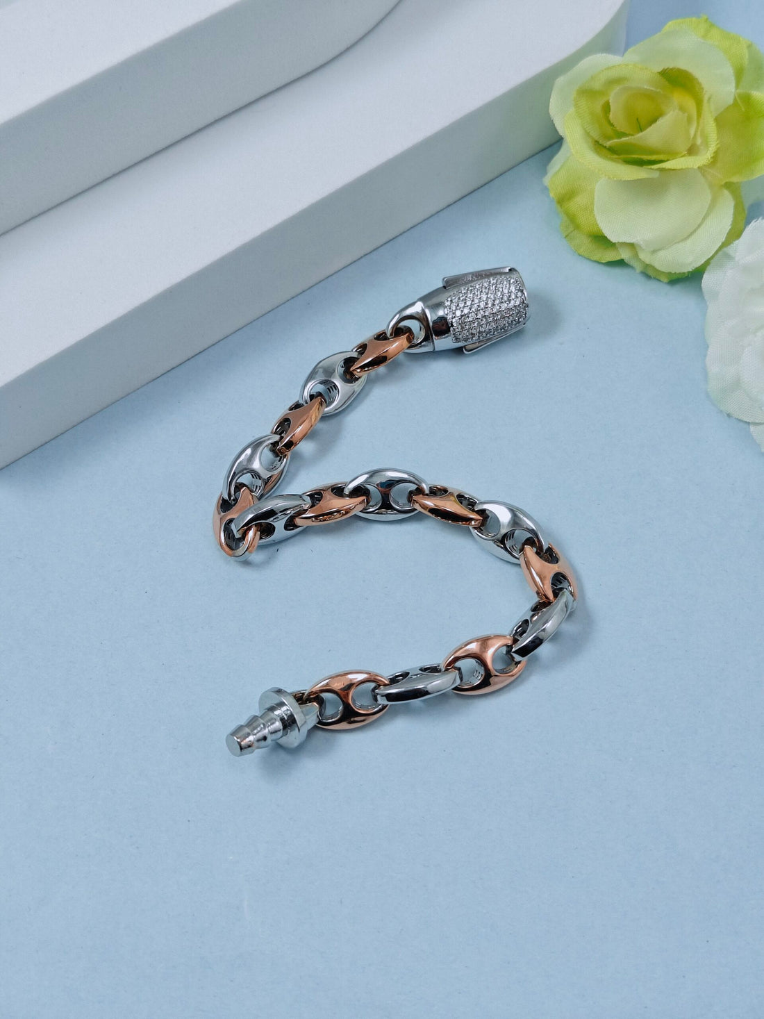 Silver and rose gold chain bracelet on a light blue surface with a white box and green flower in the background.
